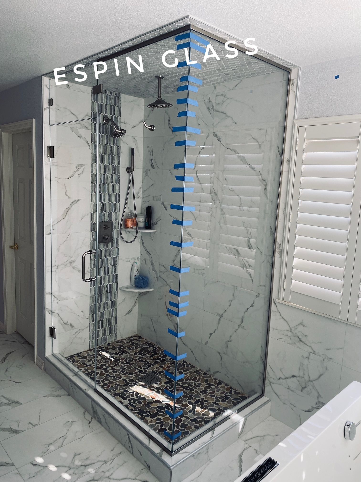 Glass shower enclosure with marble tile, pebble floor, and white shutters.