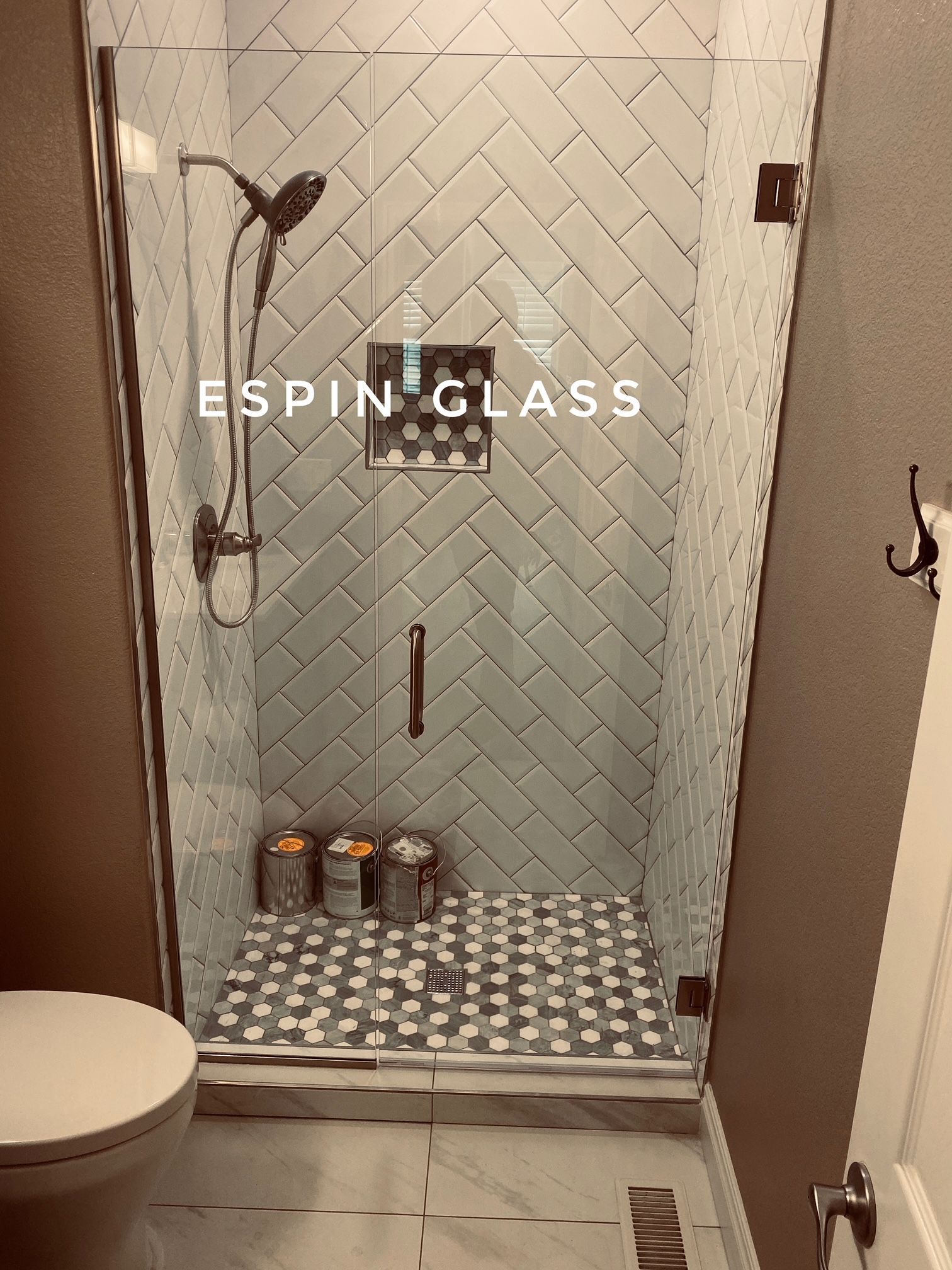 Shower with glass door, featuring white herringbone tile, and patterned floor tiles.