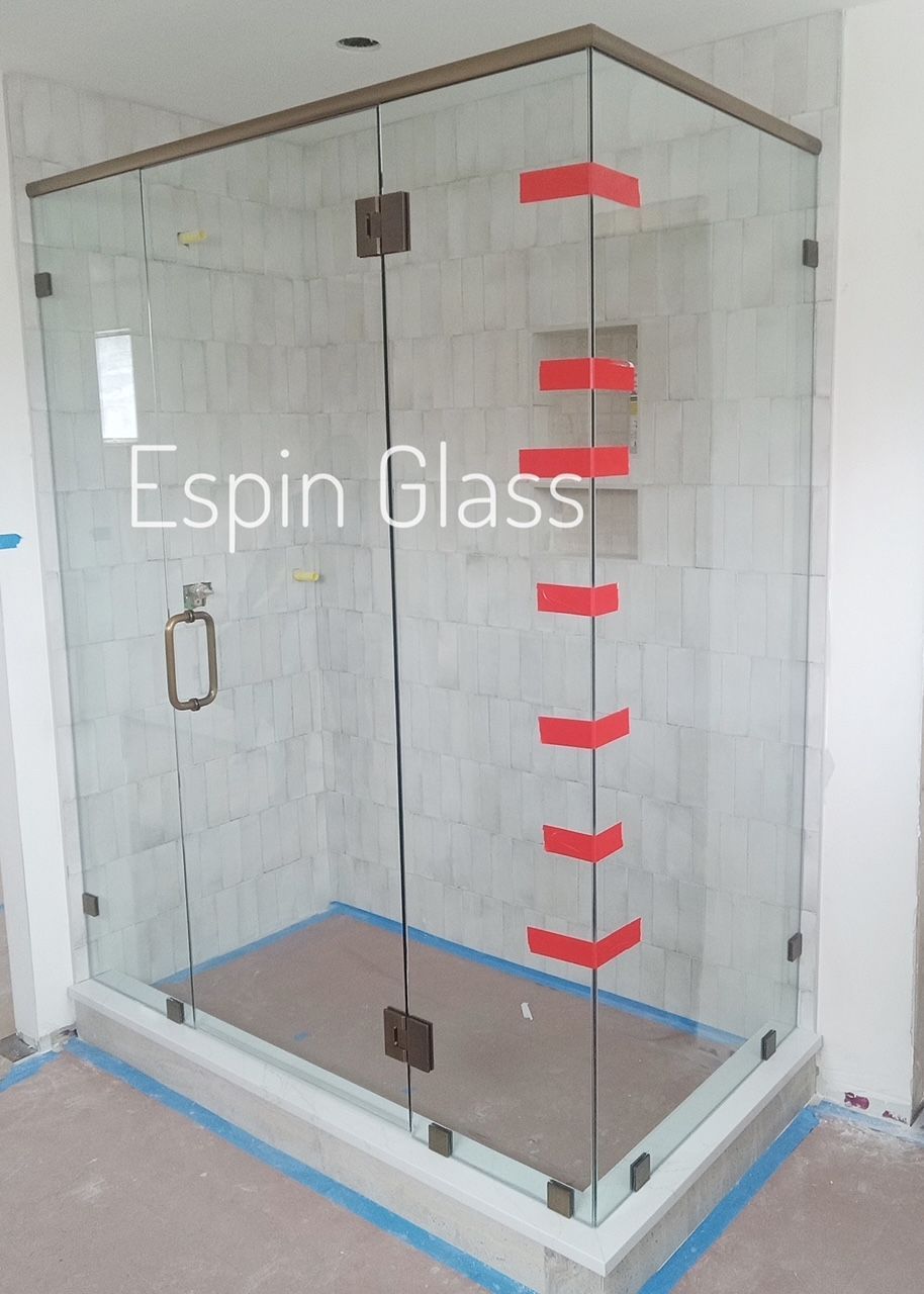 Glass shower enclosure with bronze hardware, red tape, and a light-colored tiled wall.