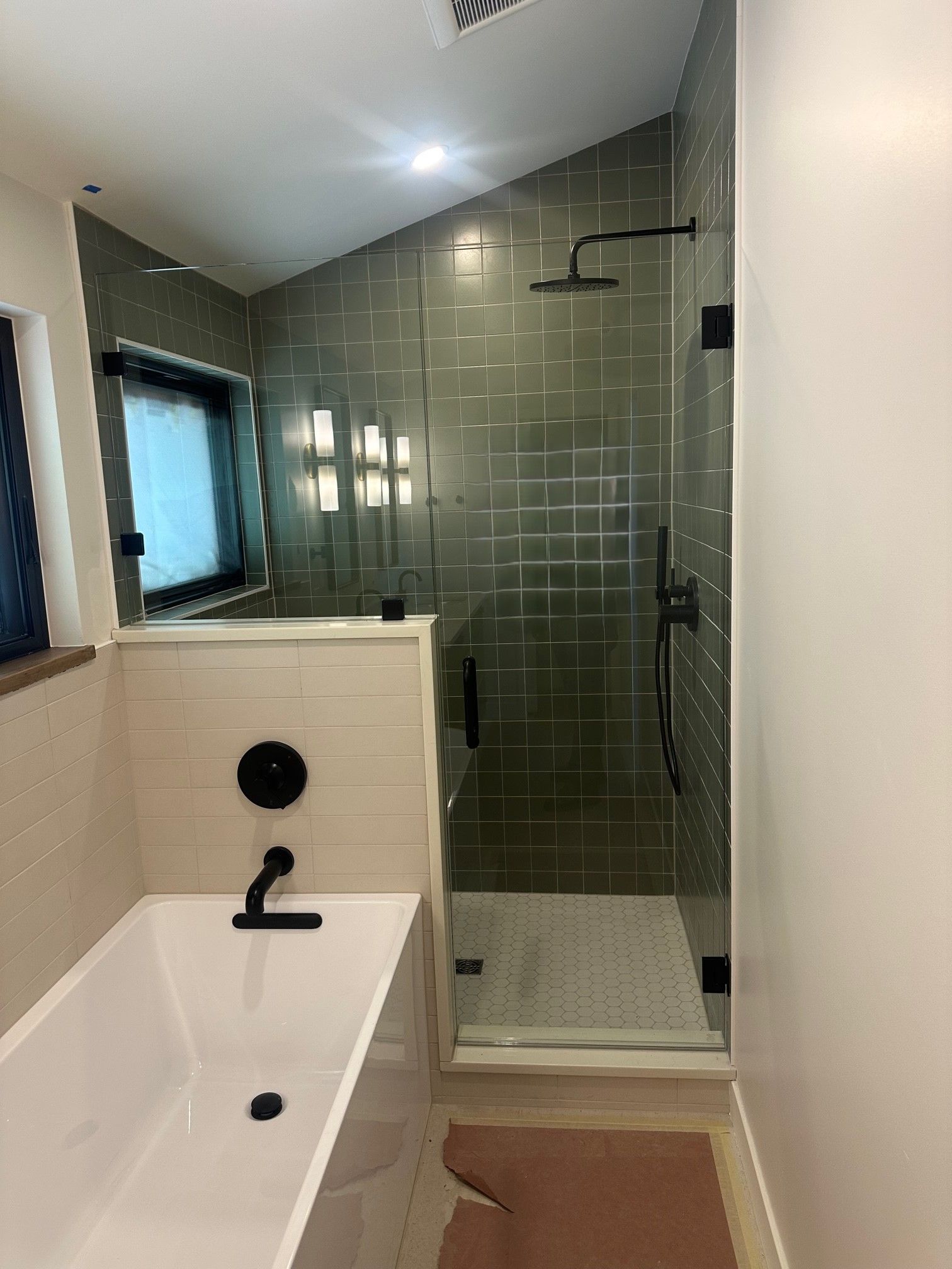 Modern bathroom with a white tub, glass shower, and black fixtures. Green tile, beige wall.