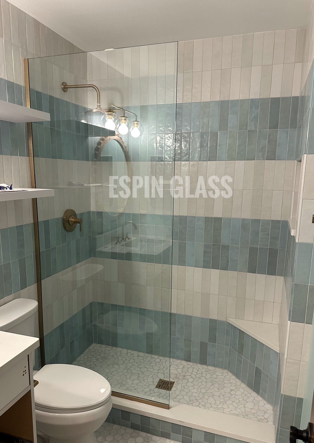 Bathroom with glass shower, blue and white tiled walls, toilet, shelves, and metal fixtures.