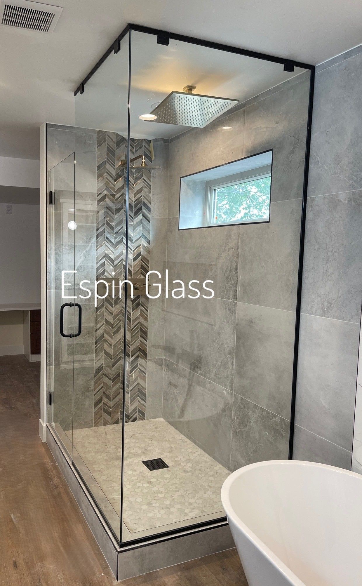 Modern glass shower with black trim, gray tile walls, and a white tub in a bathroom.
