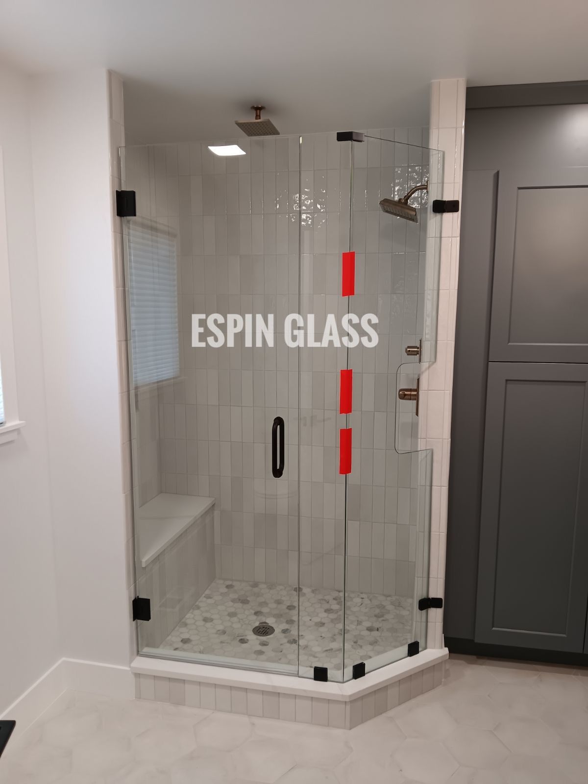 Glass shower enclosure in bathroom with light-colored tile walls and floor, black hardware, and a small bench.