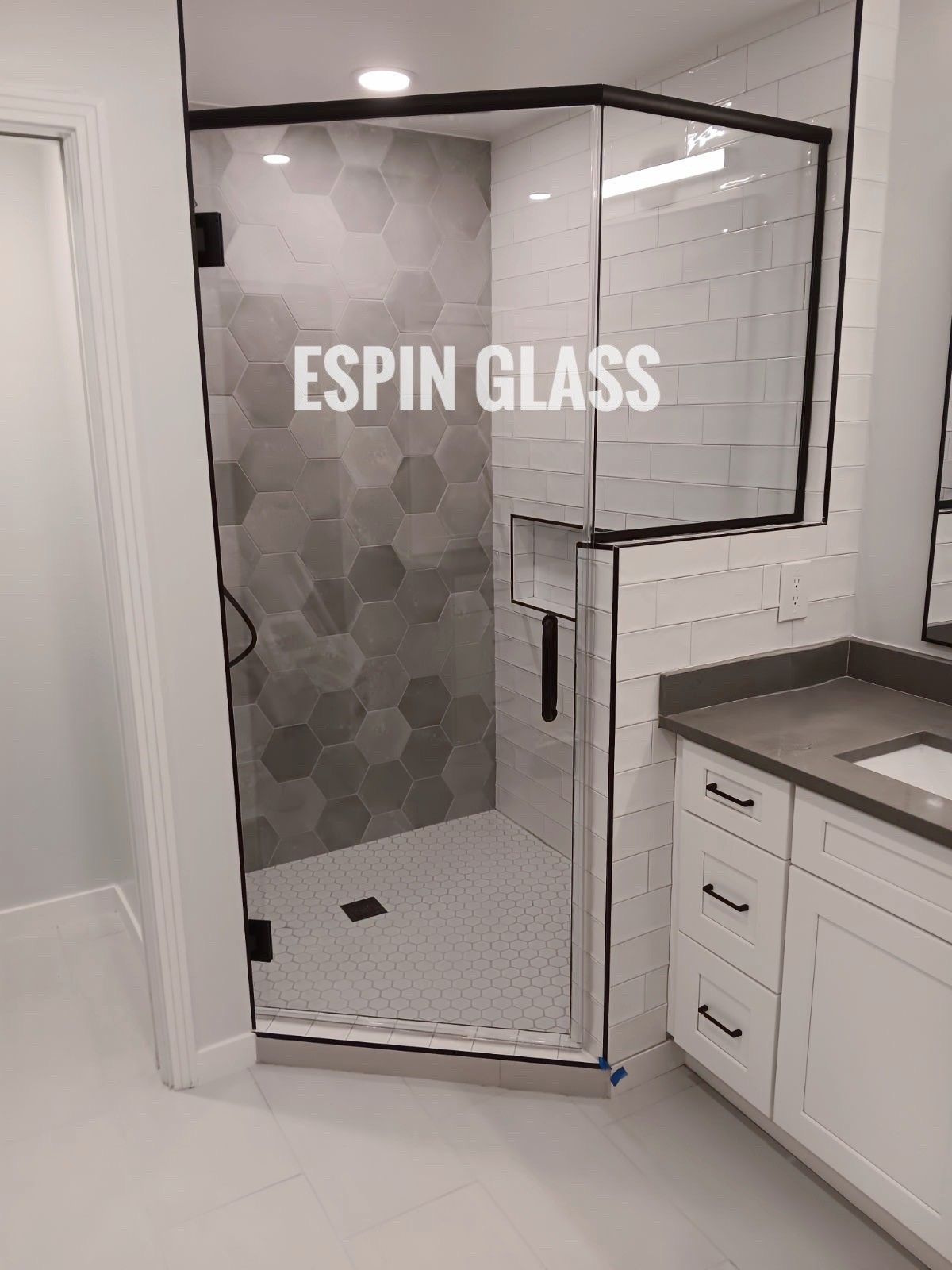 Black-framed glass shower with grey hexagon tiles, white subway tiles, and a white vanity. 