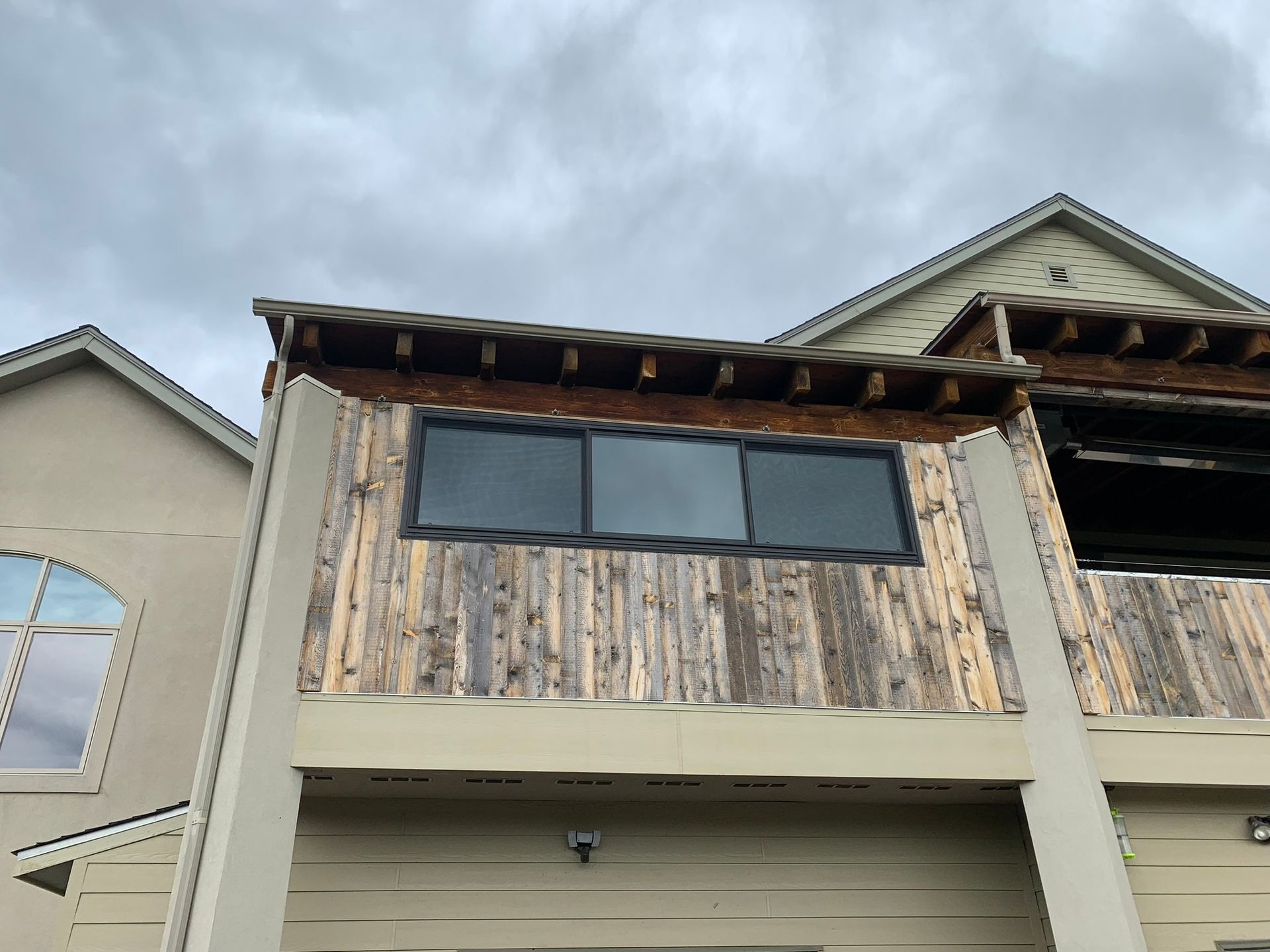 Weathered wood-sided house facade with three dark windows under a cloudy sky.