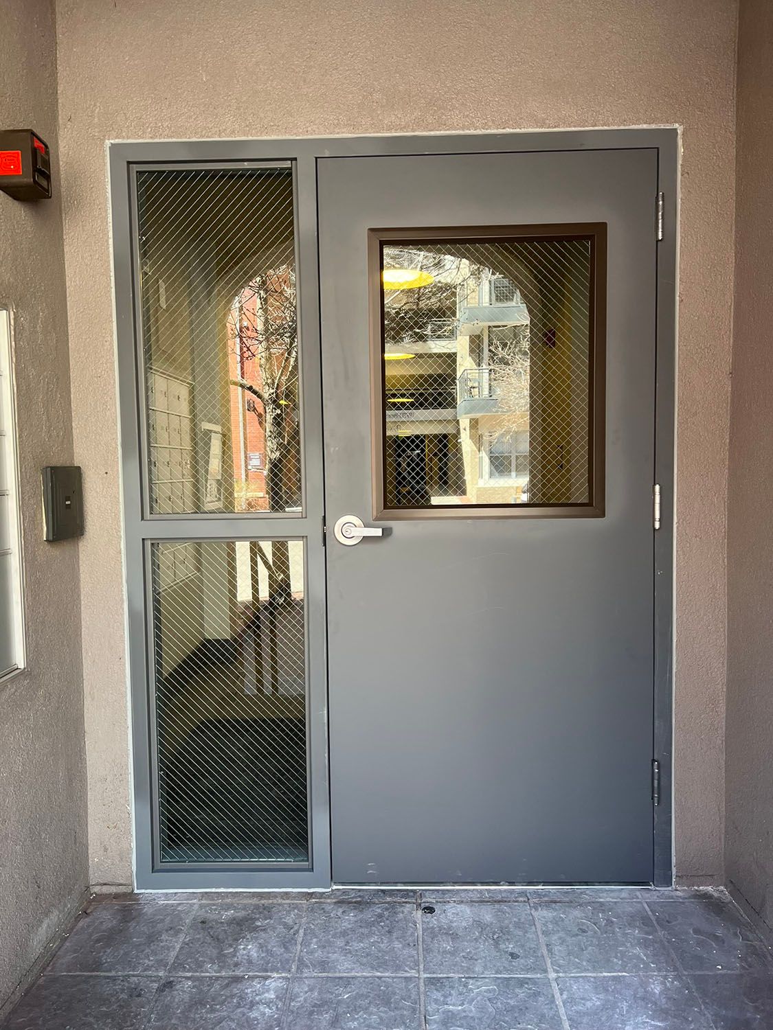 Gray exterior door with wire-glass panels and a small window, set in a stucco building entrance.