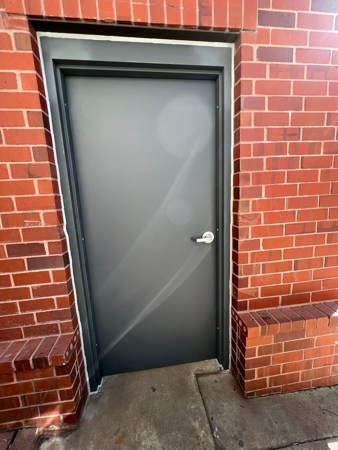 Gray metal door in a red brick wall with a silver handle and small concrete step.