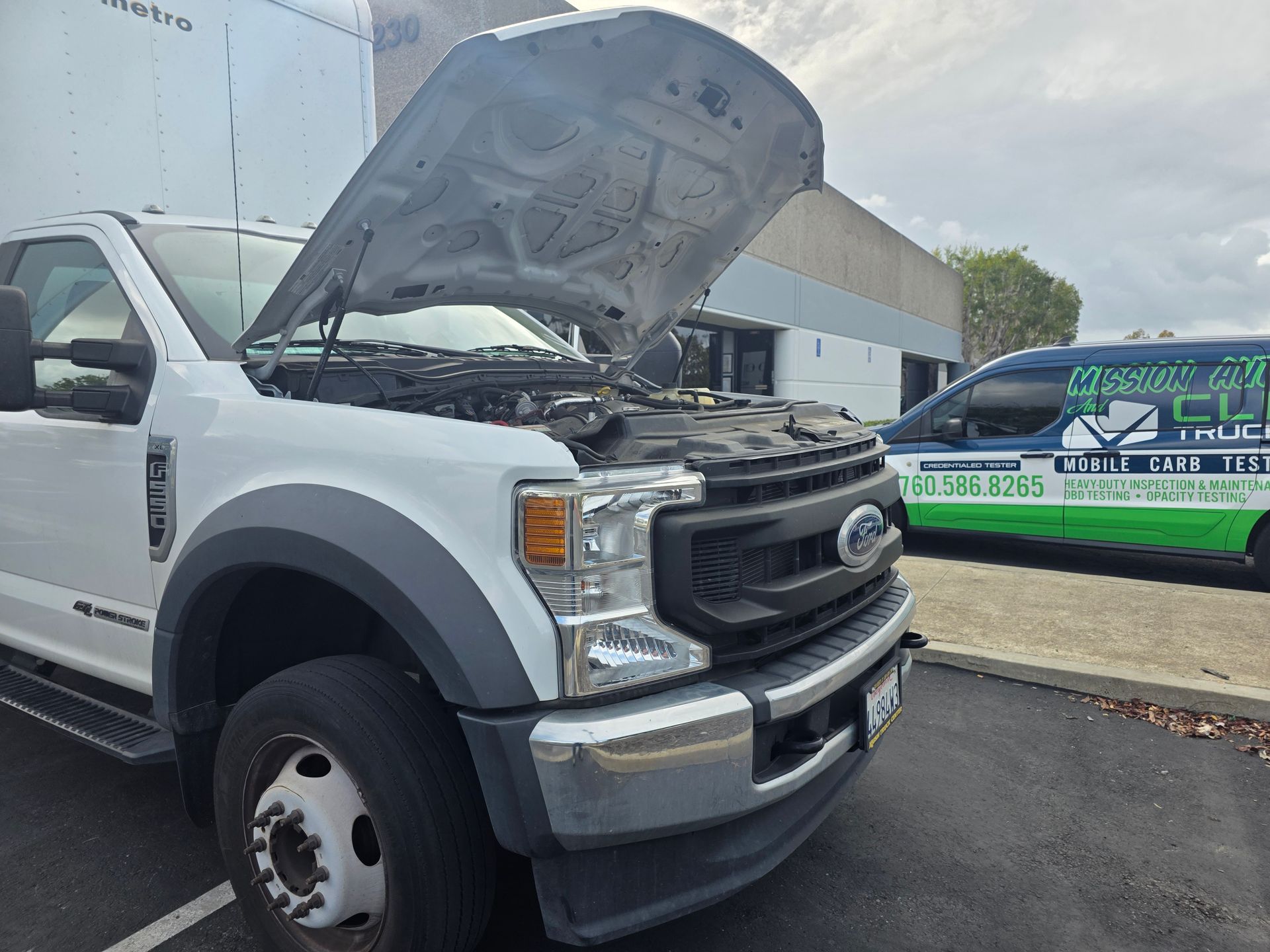White Ford truck with hood open, parked outside a garage beside a green service van.
