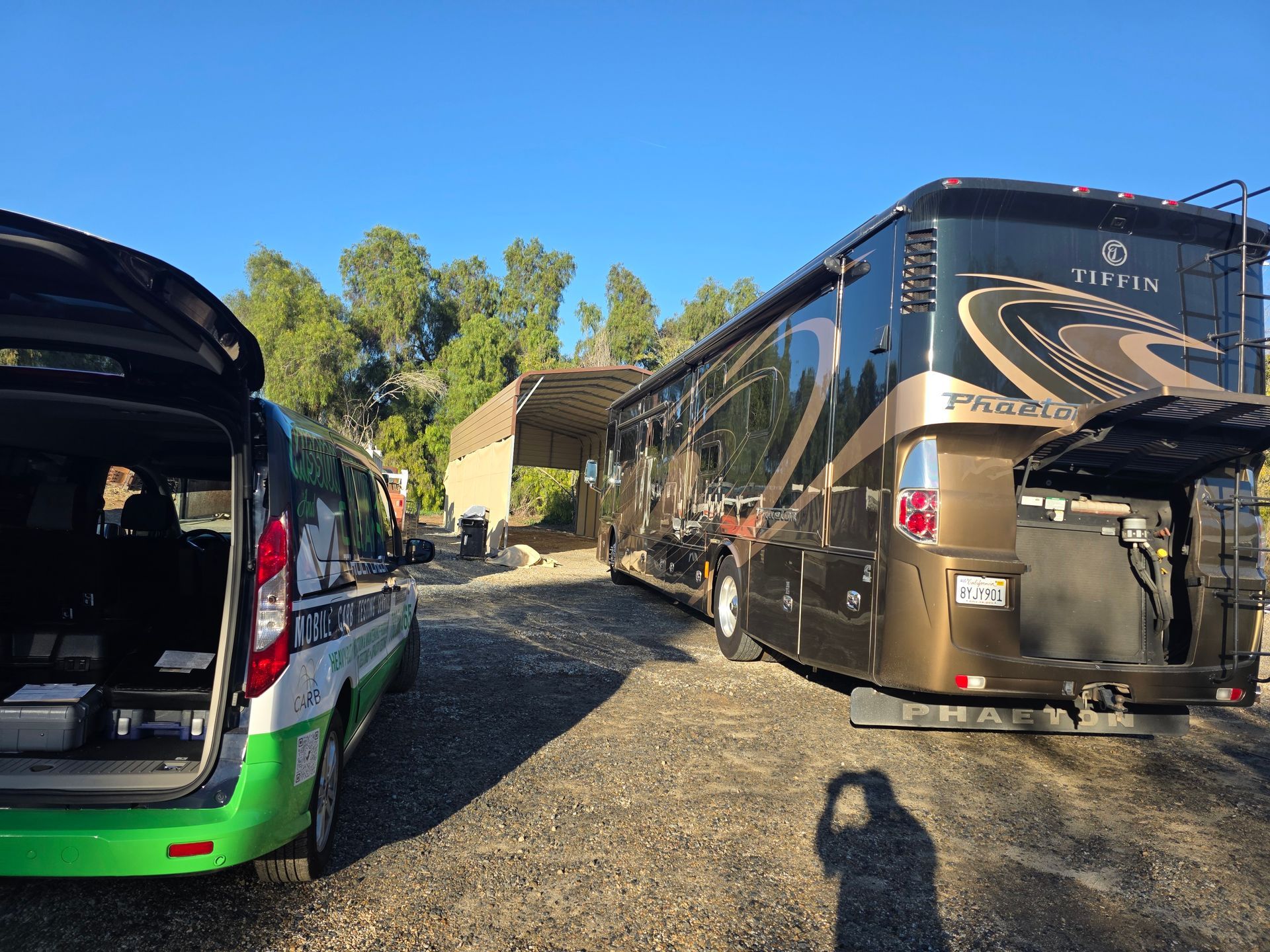 Two RVs parked on a gravel lot in bright sunlight, with trees and a building in the background.