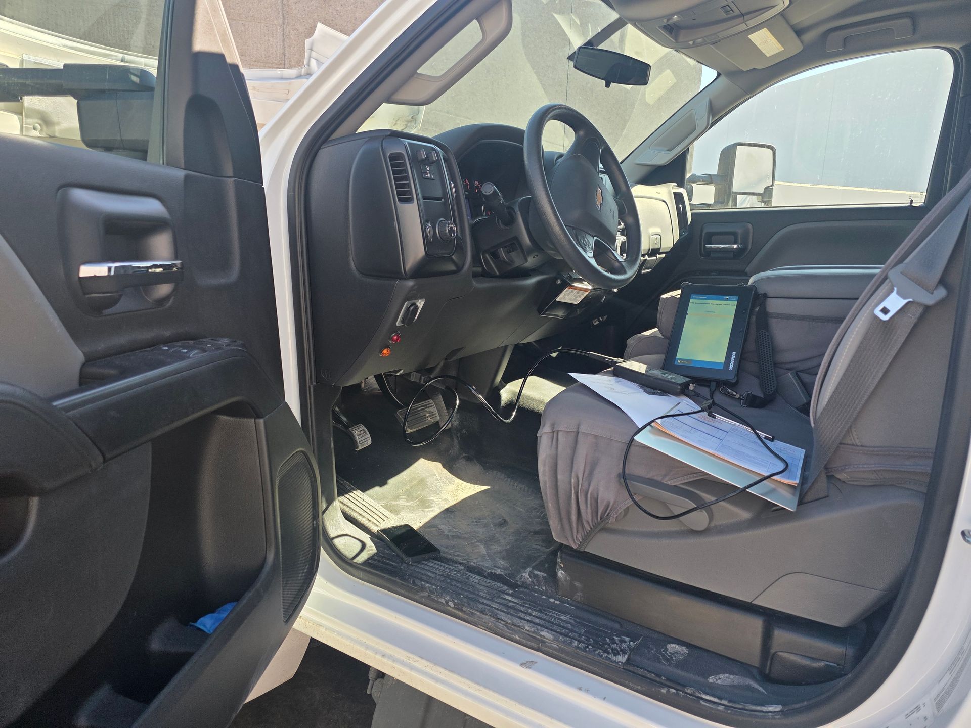 Car interior with driver door open, laptop and papers on passenger seat, wheel and dashboard visible.