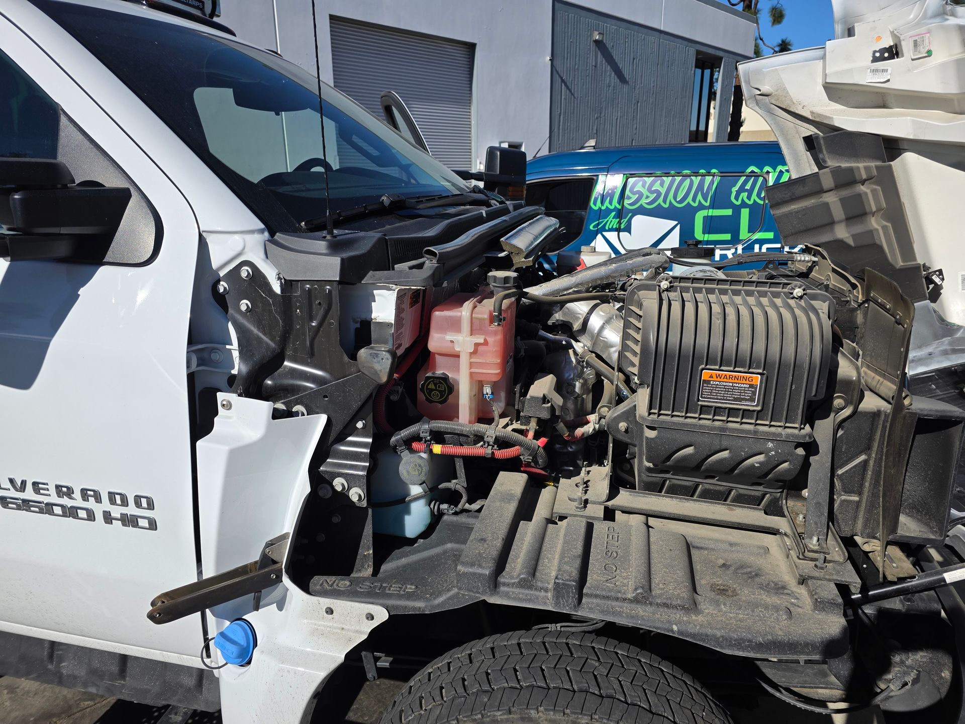 Severely damaged white truck with exposed engine after a front-end crash, parked outdoors near a tow truck
