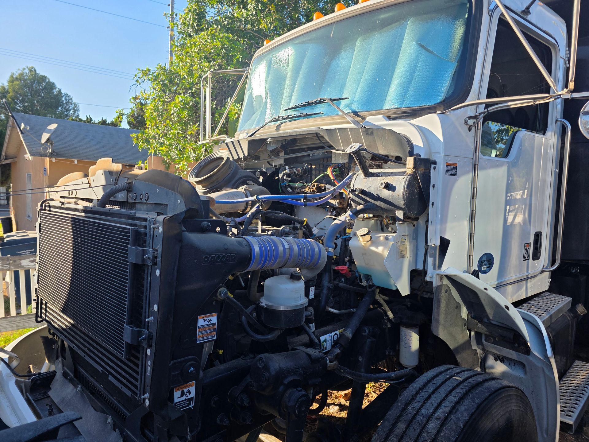 Damaged semi-truck front end with exposed engine after a collision, parked outdoors under trees