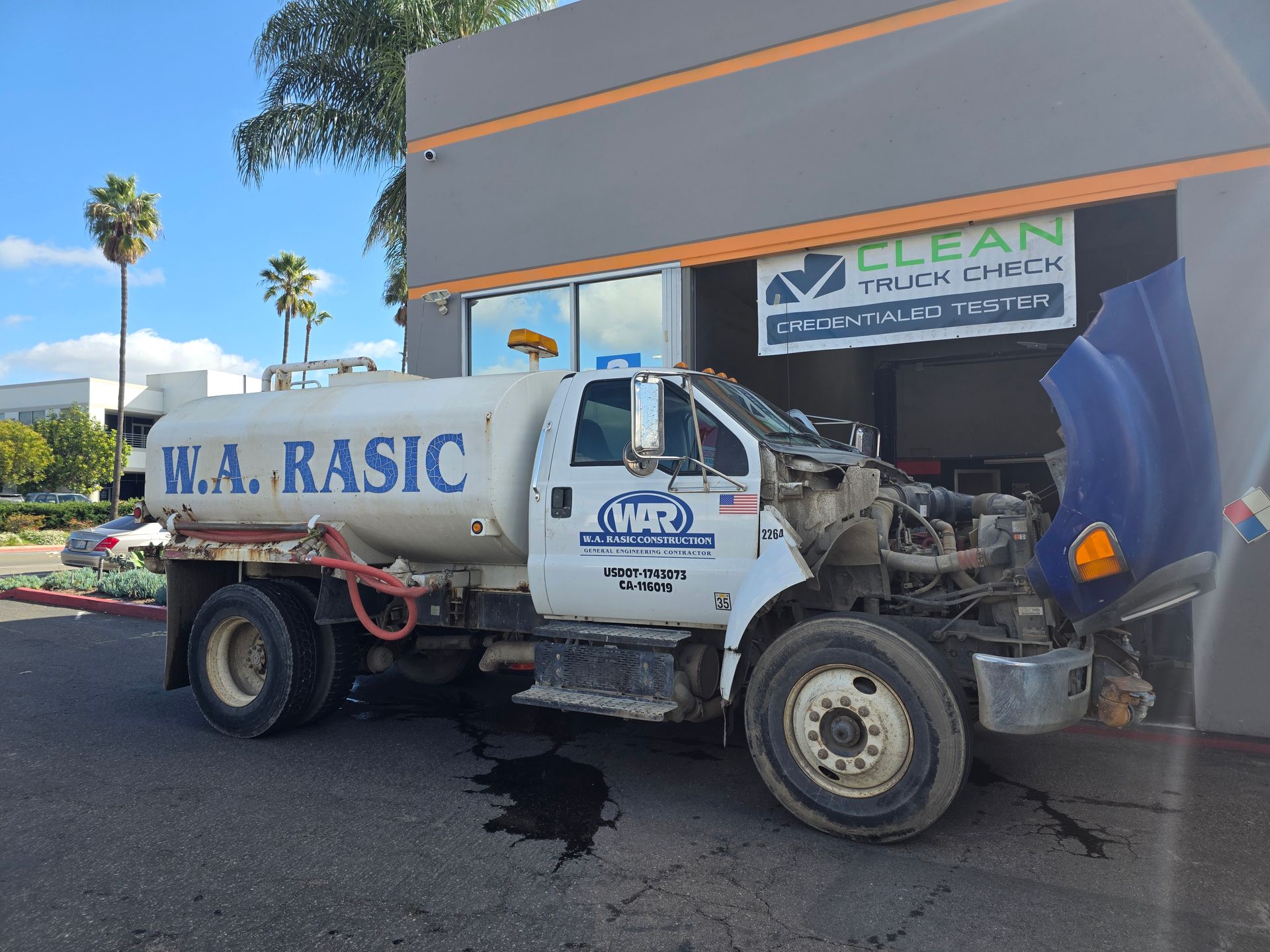 White W.A. Basic water truck crashed into a building storefront, with debris and a damaged front end.