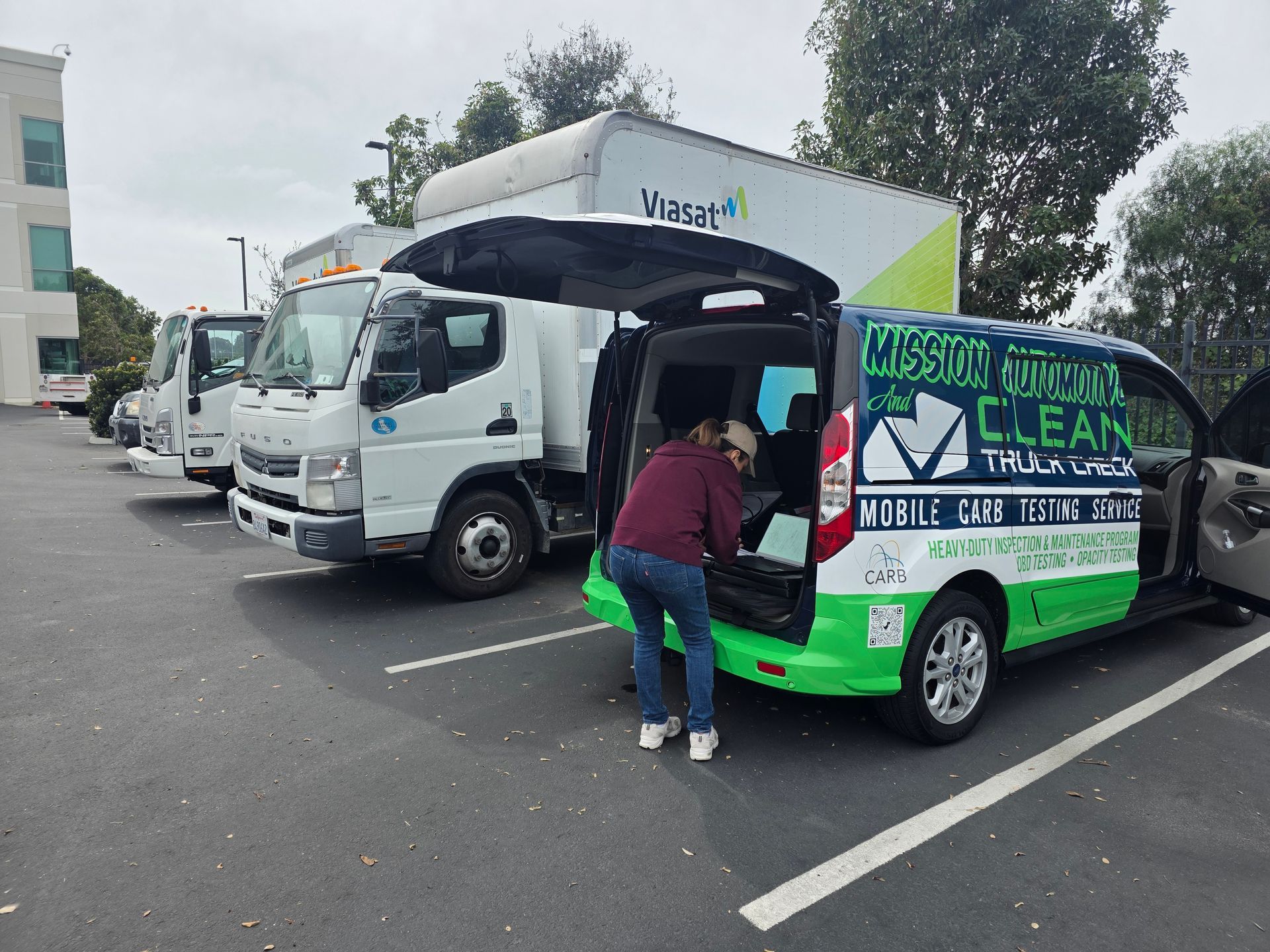 Person loading supplies into a green community outreach van beside a white box truck in a parking lot