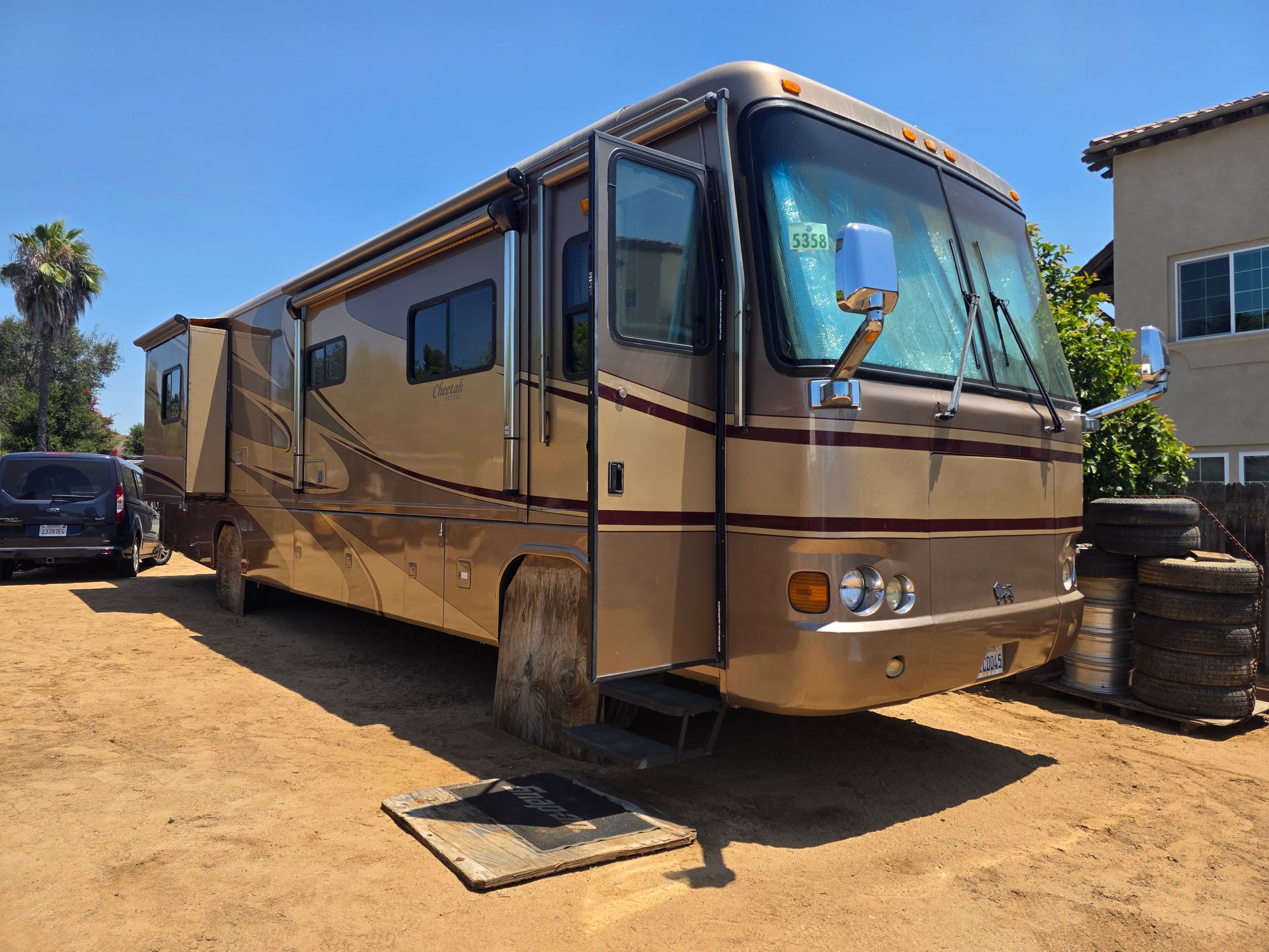 Tan motorhome parked on a dirt lot beside a building, with windows and a side awning.