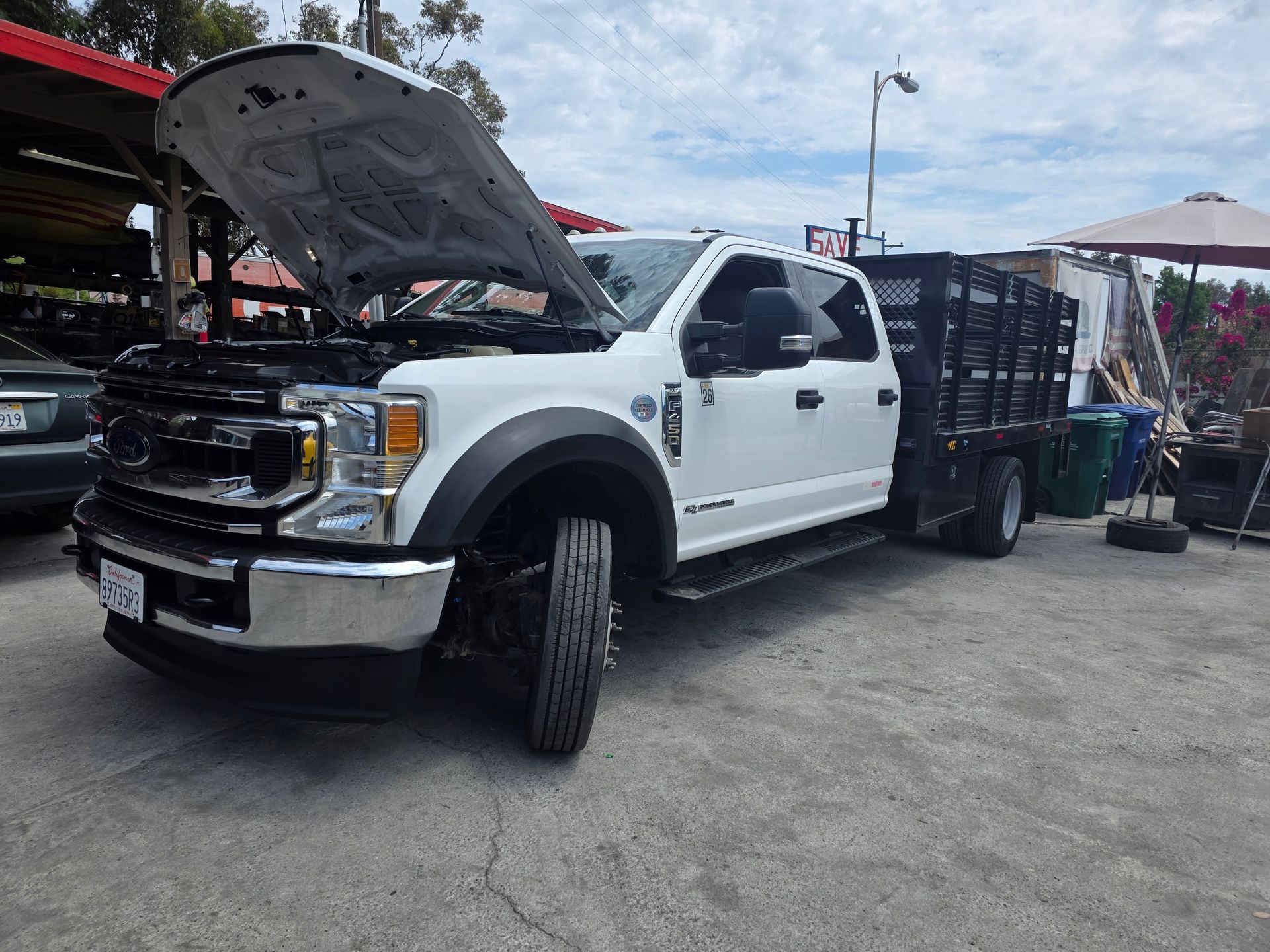White Ford pickup with hood open at an outdoor market or car show, parked beside vendor tents and displays