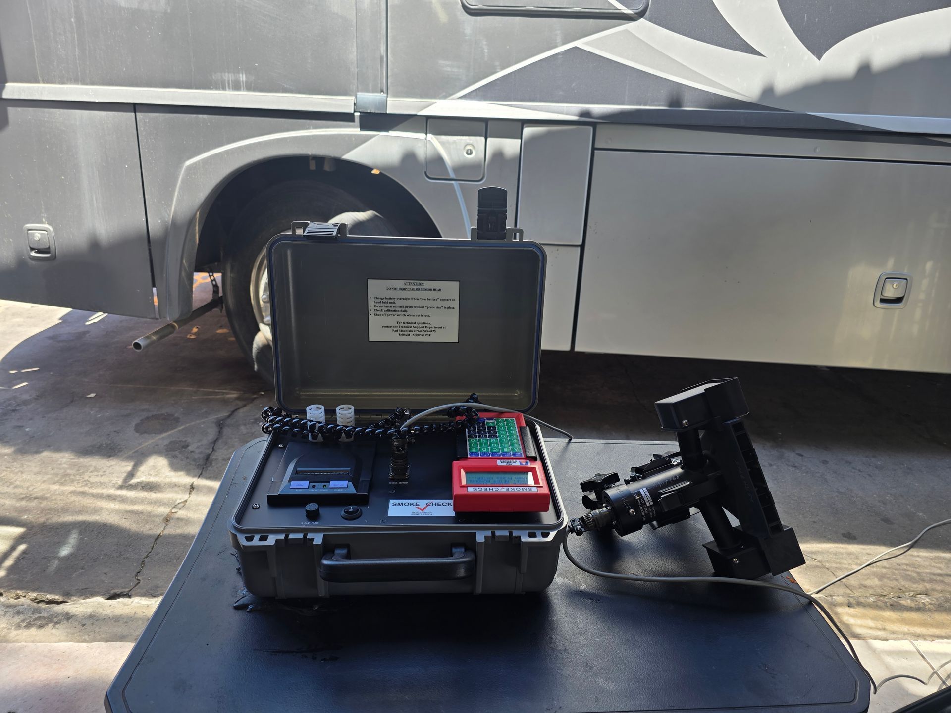 Portable electronic testing rig with open black case and mounted display beside a vehicle wheel.