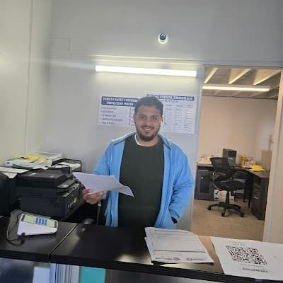 Man standing behind a counter in a shop, holding papers and smiling; printers and desks in the background