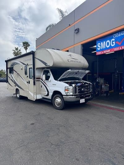 White RV parked outside a smog shop under an awning, with front cab-over cover raised.