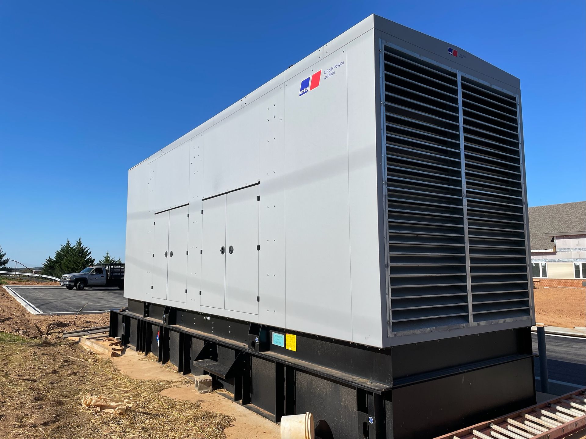 Large white industrial generator on a black base, outdoors on a dirt lot under a blue sky.