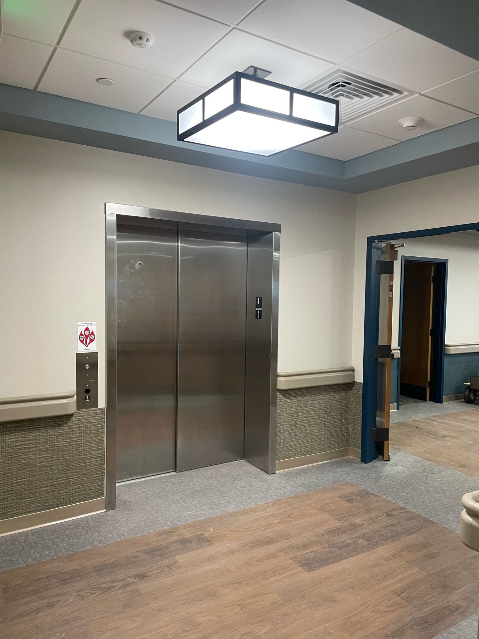 Stainless steel elevator in a hallway with wood flooring and a lit ceiling fixture