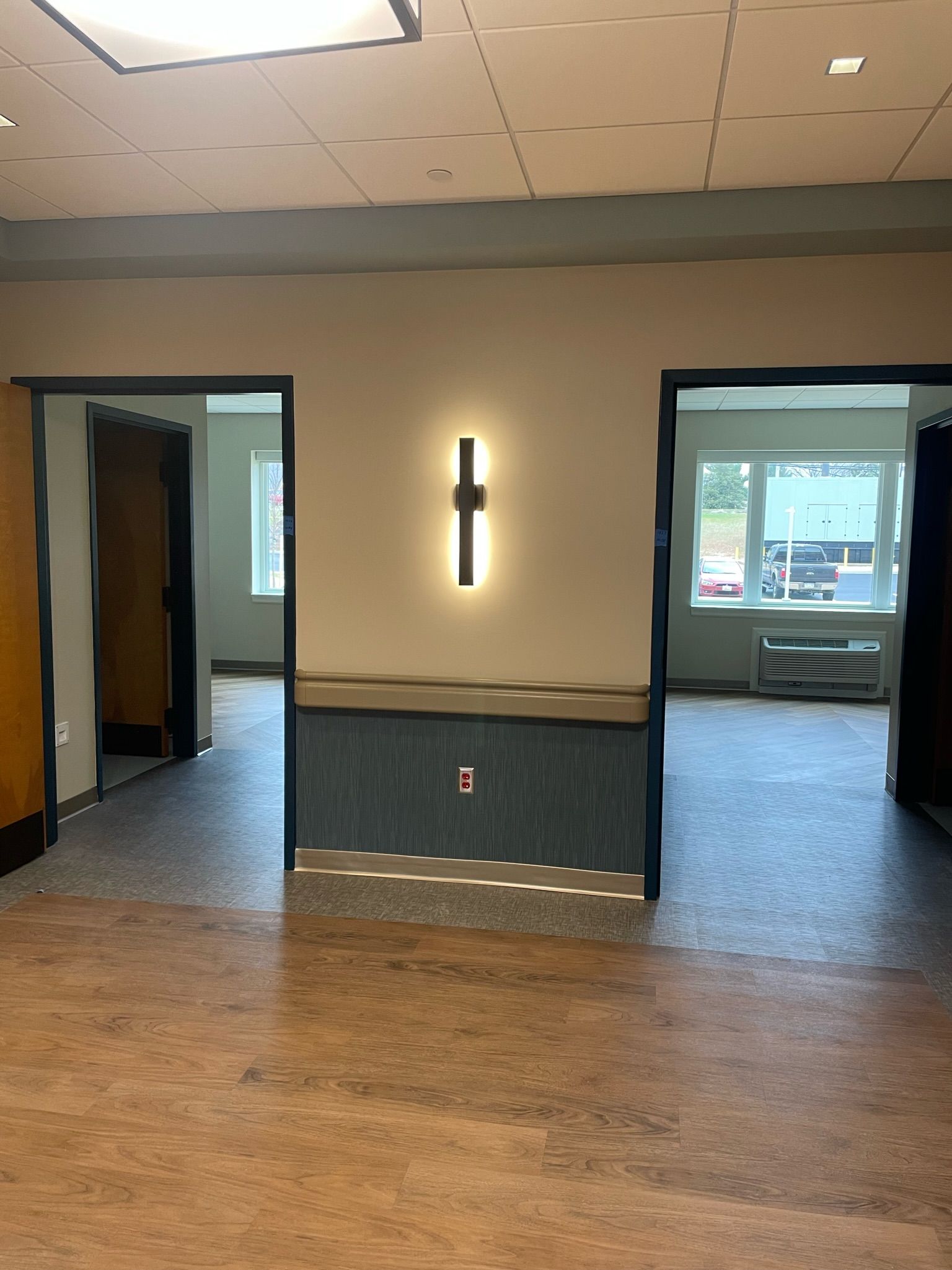 Empty lobby with wood floor, two open doorways, and a wall sconce on a beige and teal wall.