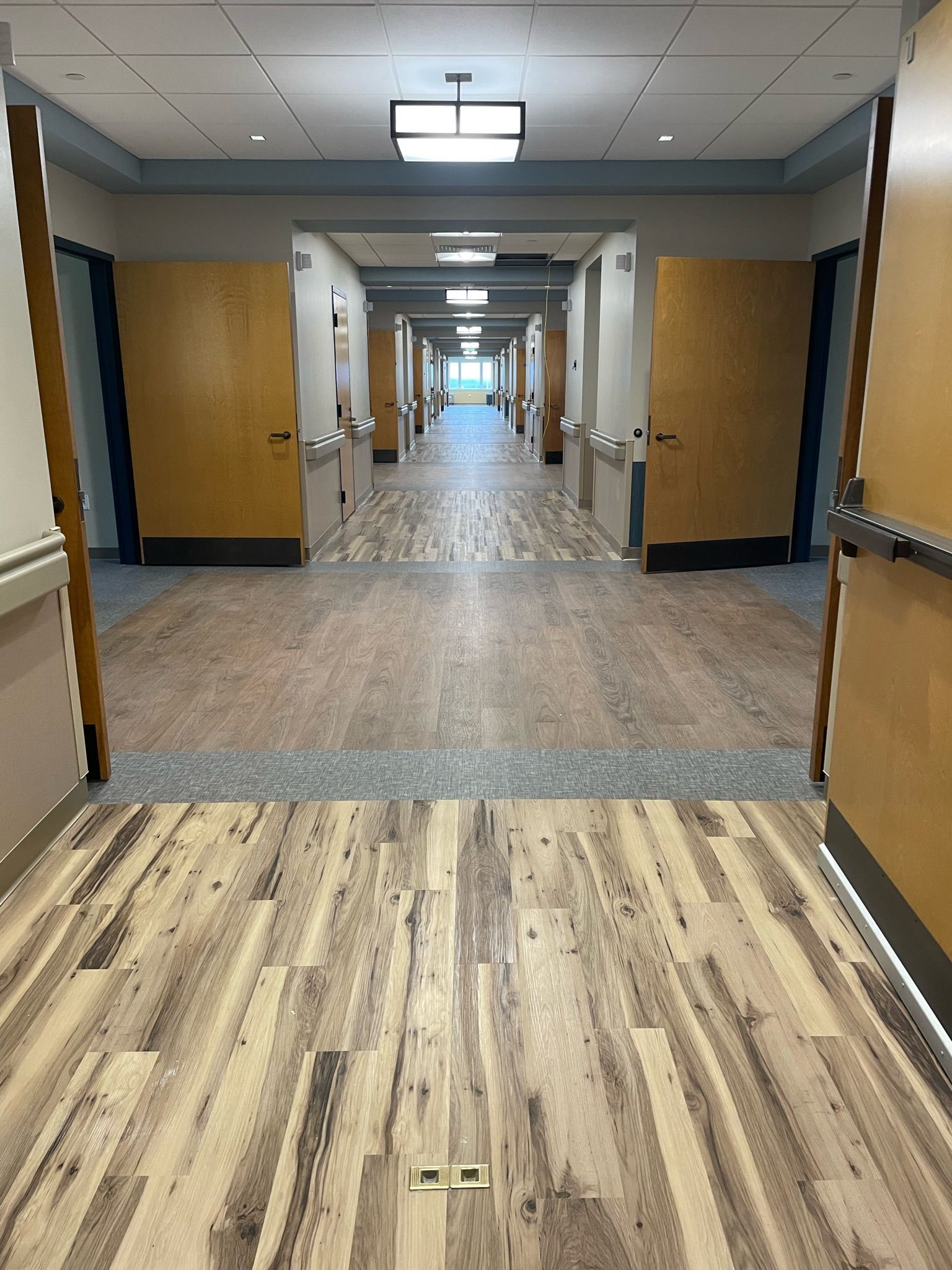 Hospital hallway with wood floor, open doors, and handrails leading to a bright corridor.
