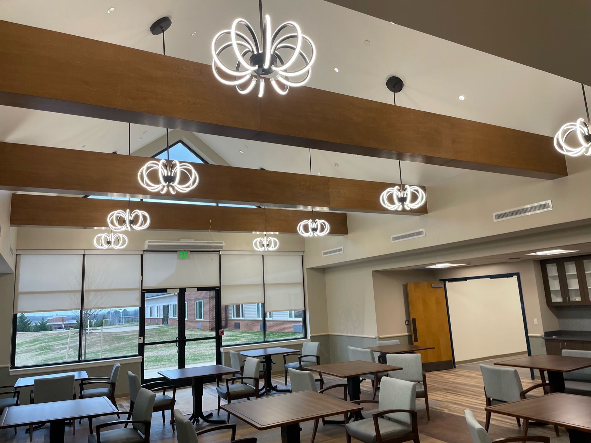 Empty classroom with tables and chairs, large windows, wood-beam ceiling, and modern pendant lights.