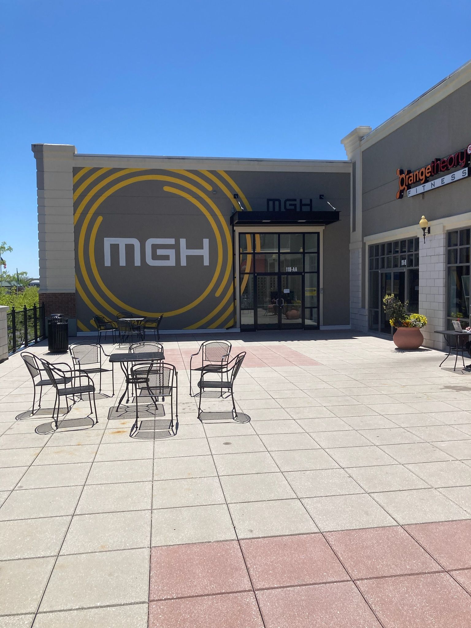 Outdoor café patio with tables and chairs in front of a building marked “MGH” under a clear blue sky