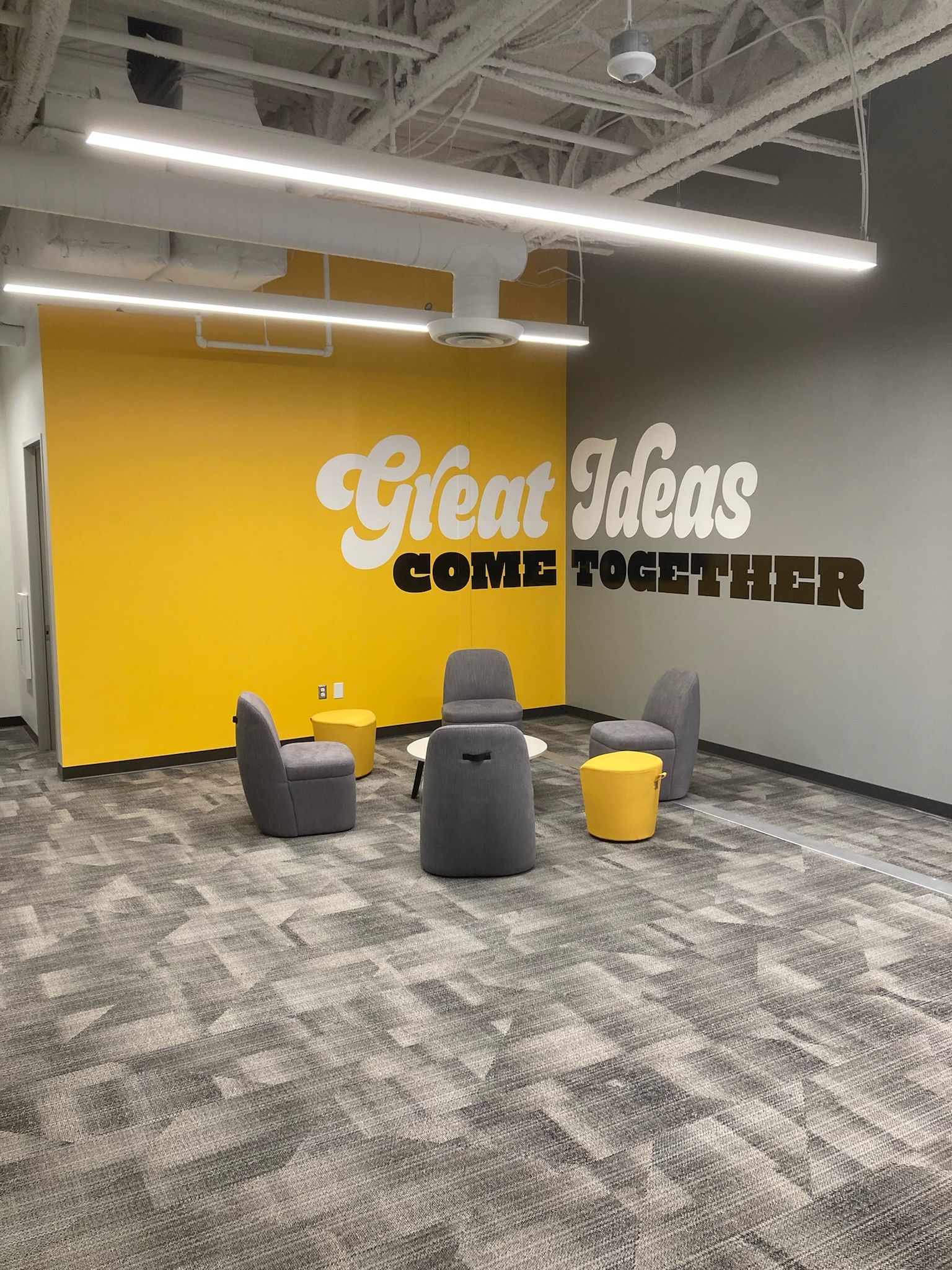 Office lounge with yellow wall reading “Great Ideas Come Together” and gray/yellow seating in an open workspace