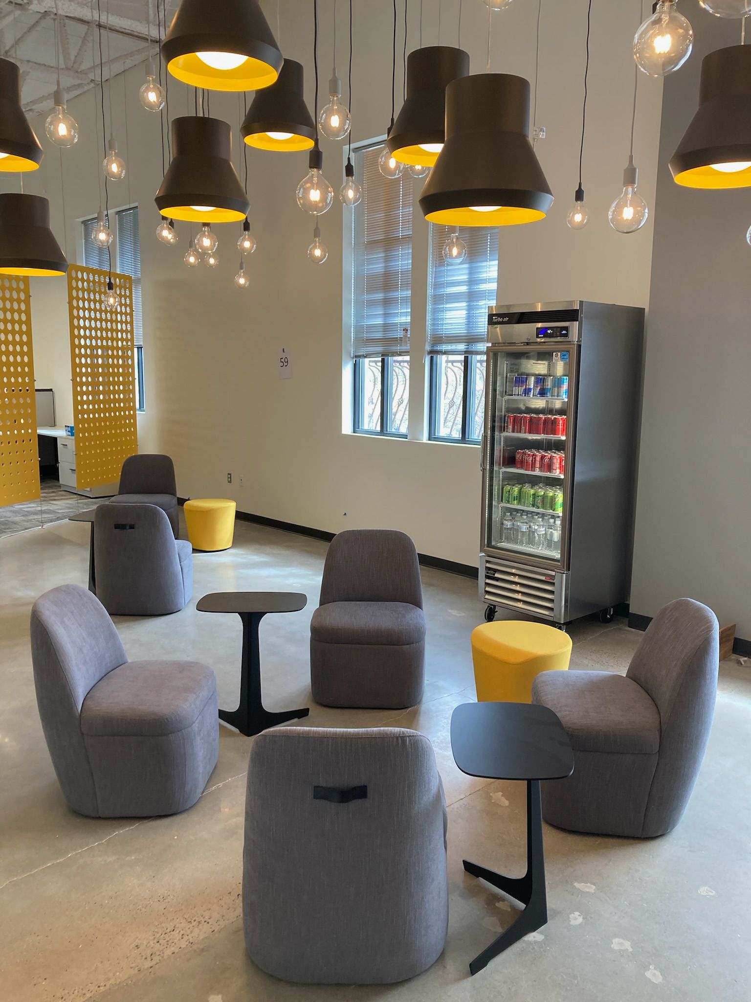 Modern lounge with gray chairs, black tables, yellow pendant lights, and a drink fridge by the windows