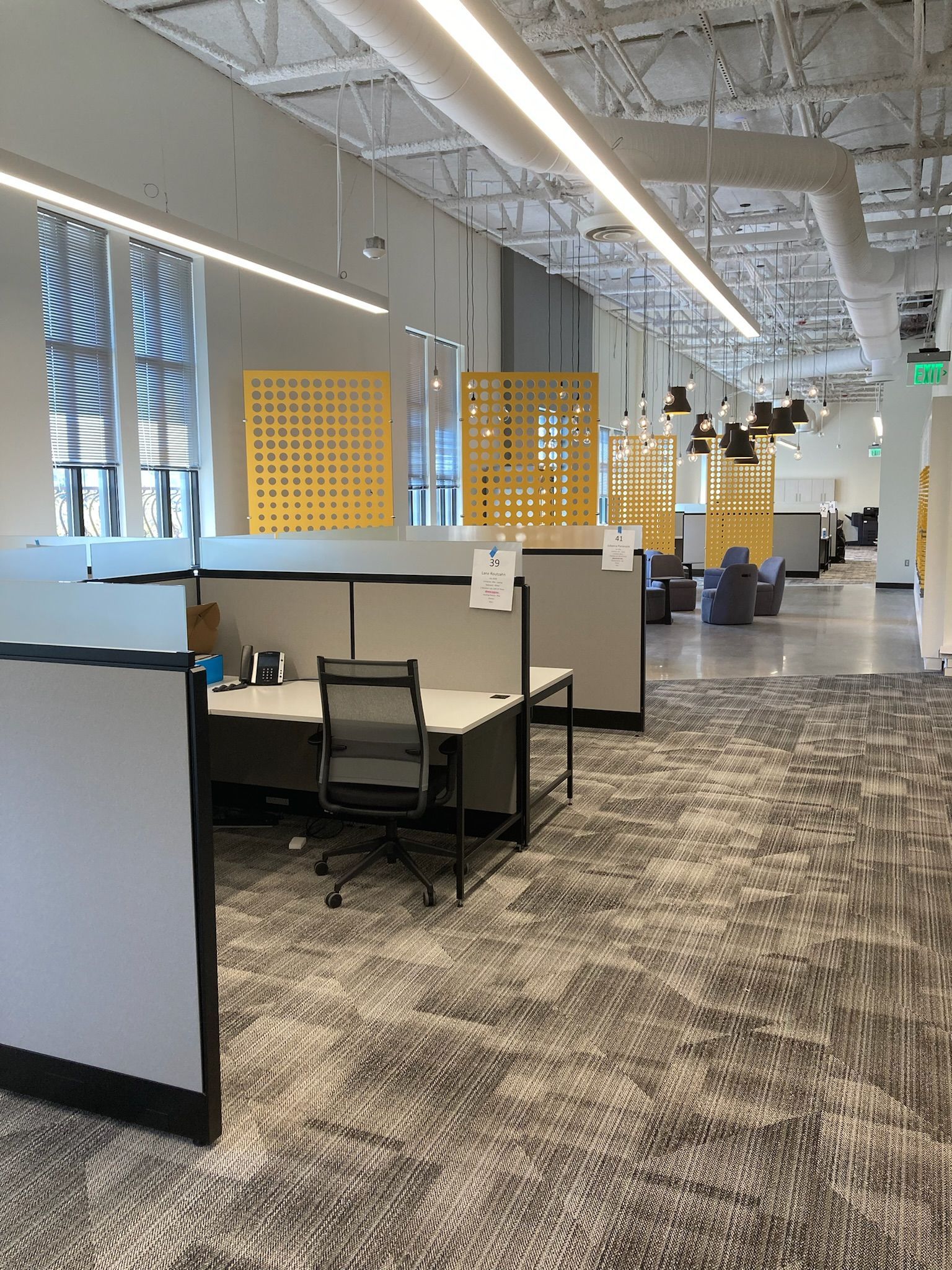 Open office workspace with cubicles, black chairs, yellow dividers, and exposed ceiling lights