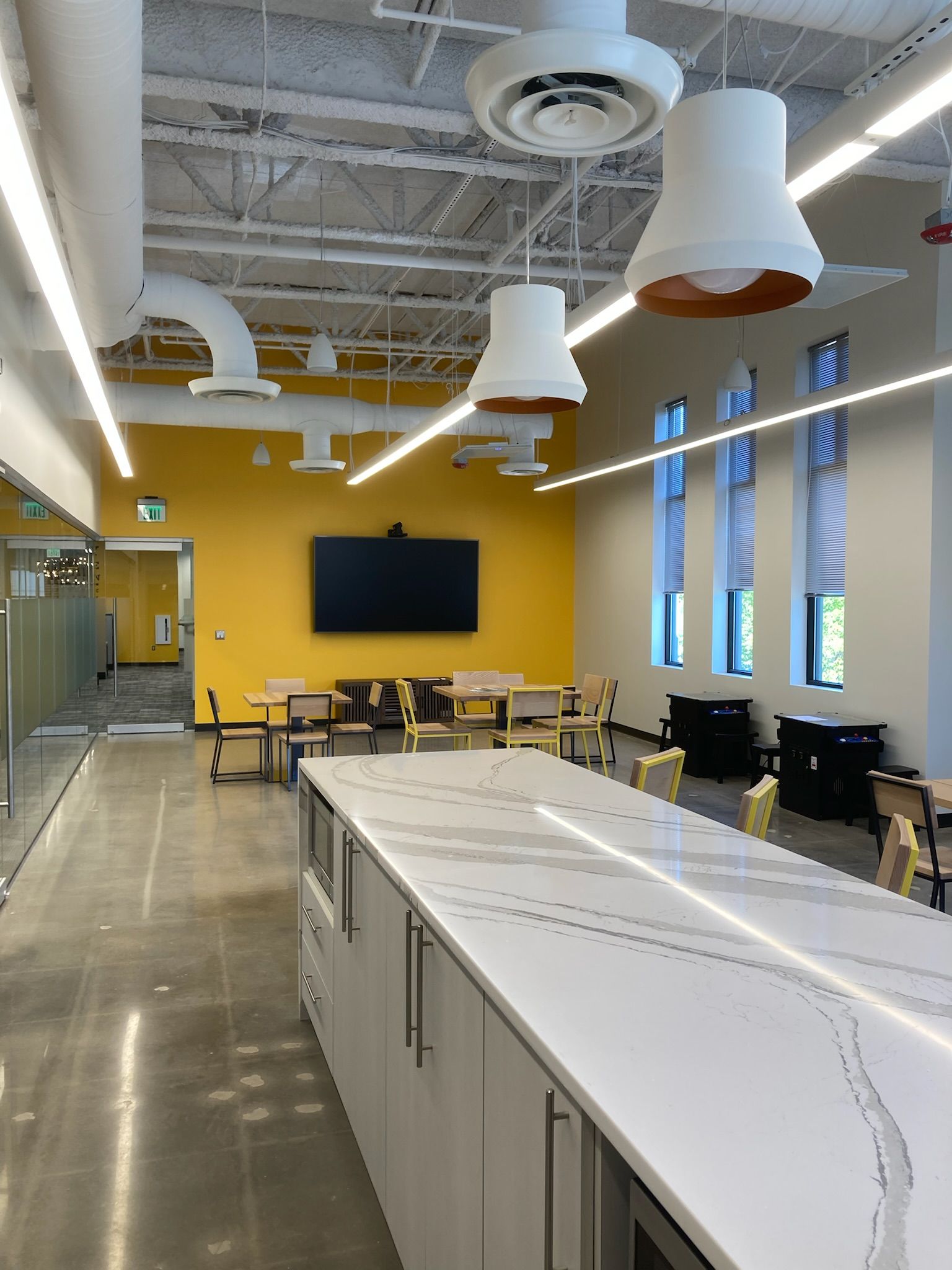 Modern cafeteria with yellow walls, white pendant lights, long tables, and a kitchen counter in the foreground