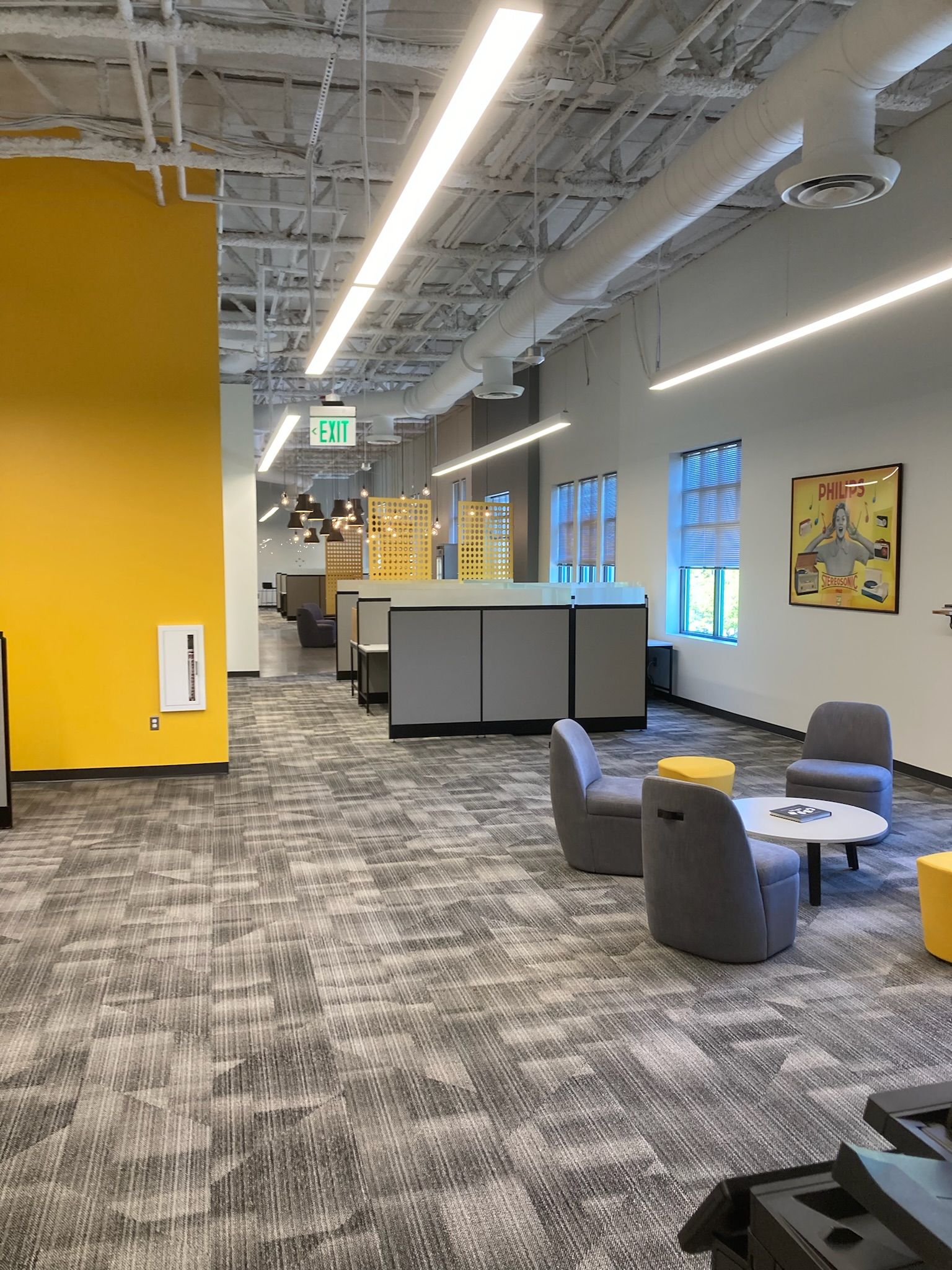 Modern open office with gray carpet, yellow wall, cubicles, lounge chairs, and large windows