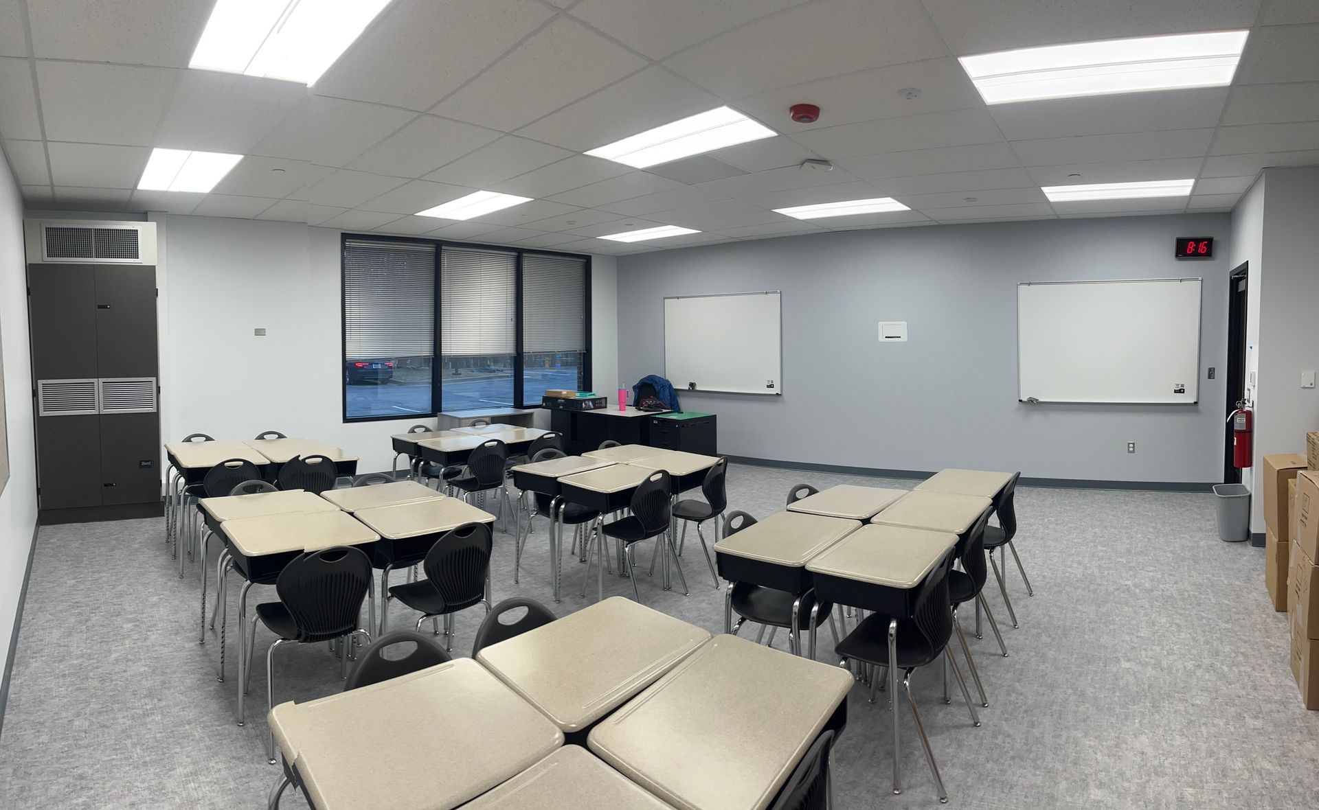 Empty classroom with beige desks, whiteboards, gray walls, and bright ceiling lights.