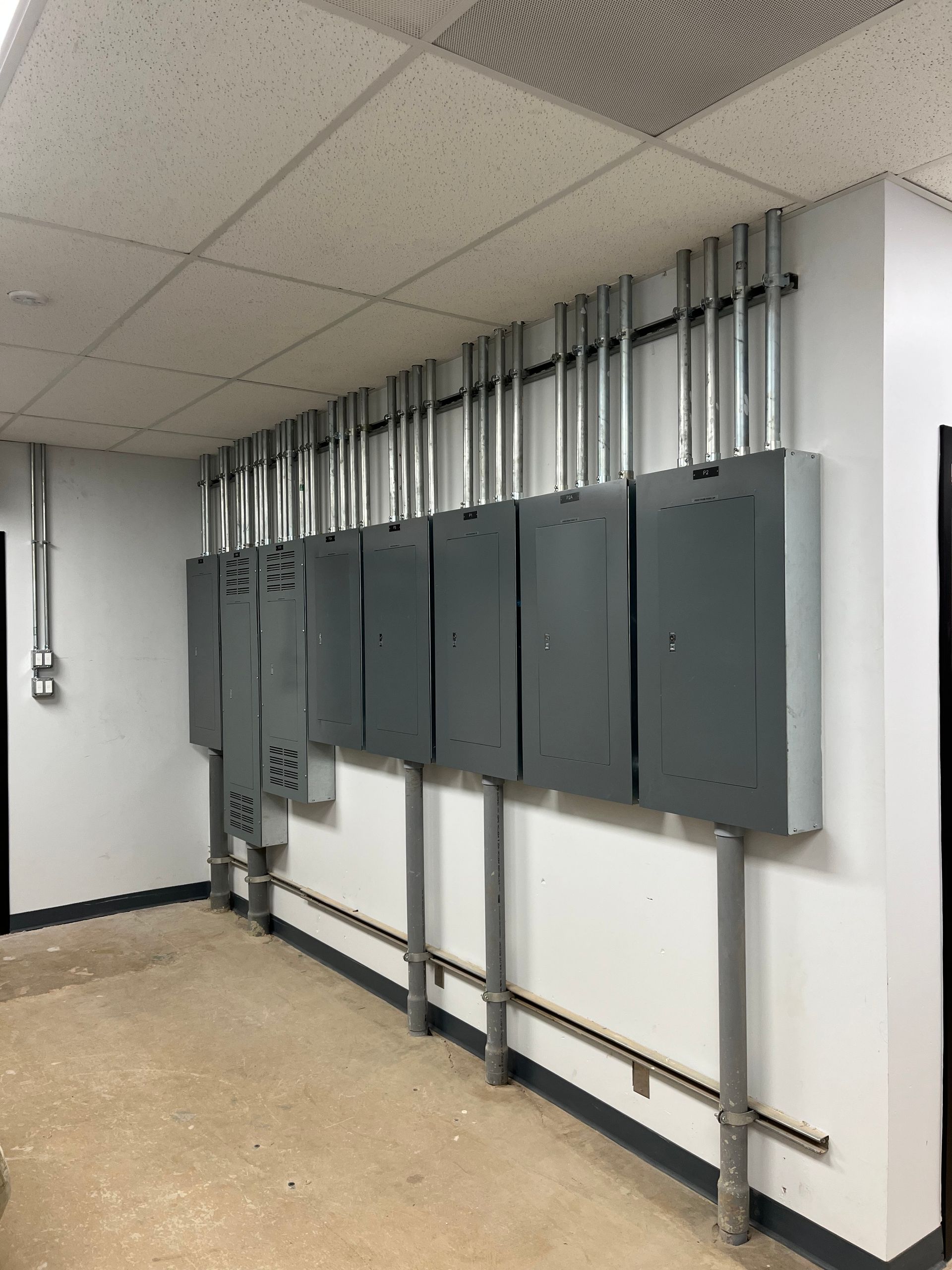 Gray electrical panels on a white wall in a utility room with exposed conduits and a concrete floor