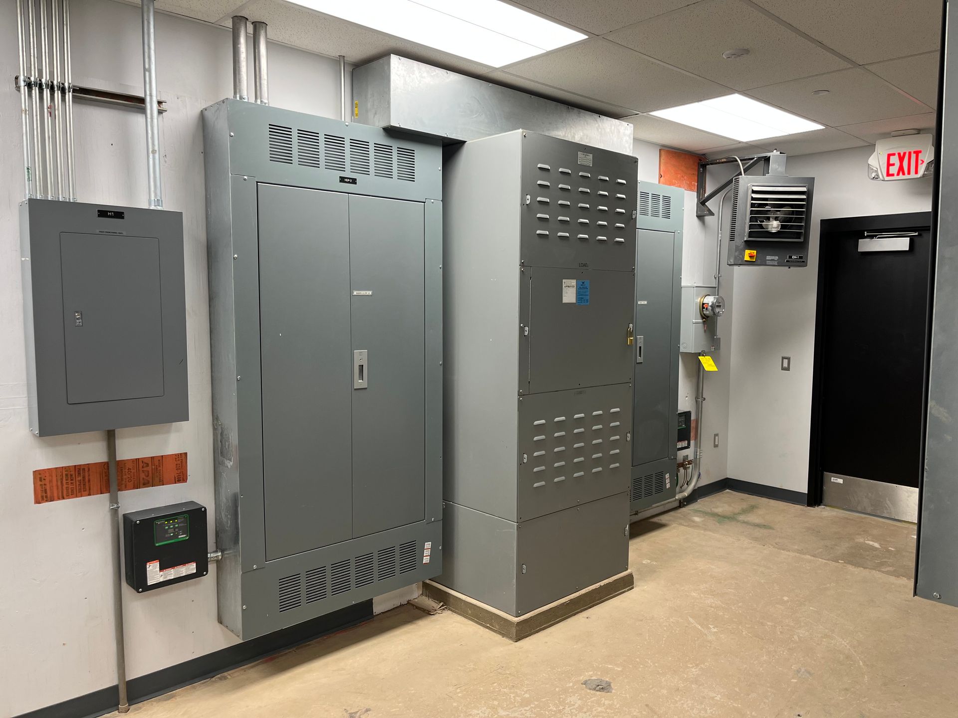 Electrical room with gray metal panels, conduit, and a black door in a hallway