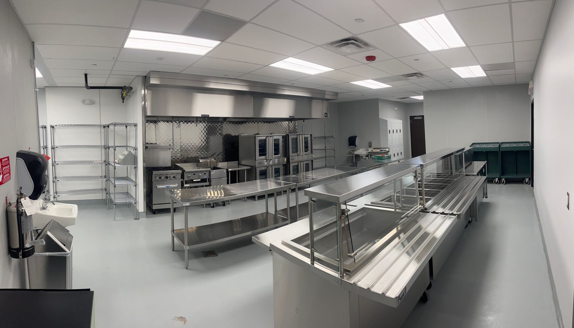 Stainless steel food prep kitchen with counters, sinks, shelves, and bright overhead lights