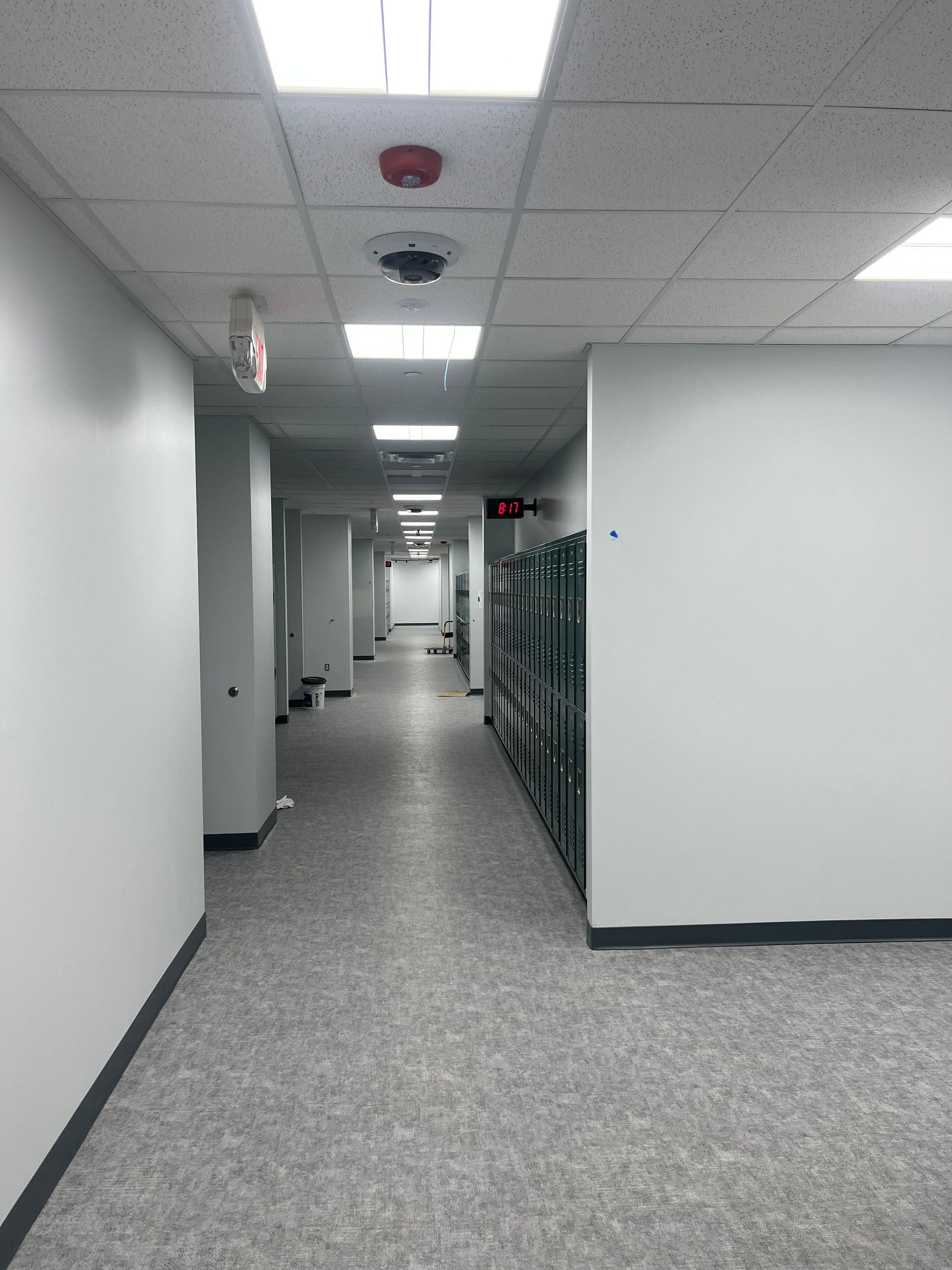 Empty office hallway with gray carpet, white walls, fluorescent ceiling lights, and lockers on the right.
