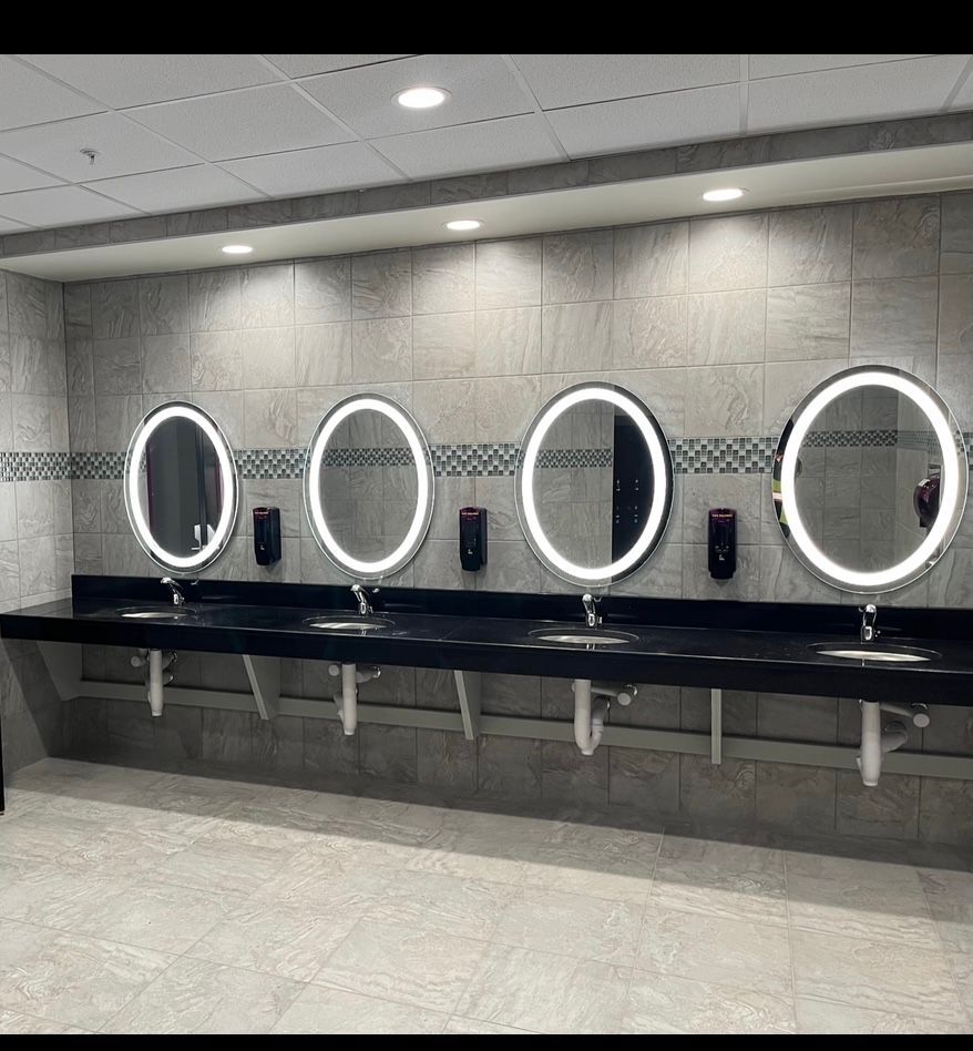 Modern restroom with four wall-mounted sinks and round backlit mirrors on a gray tiled wall.