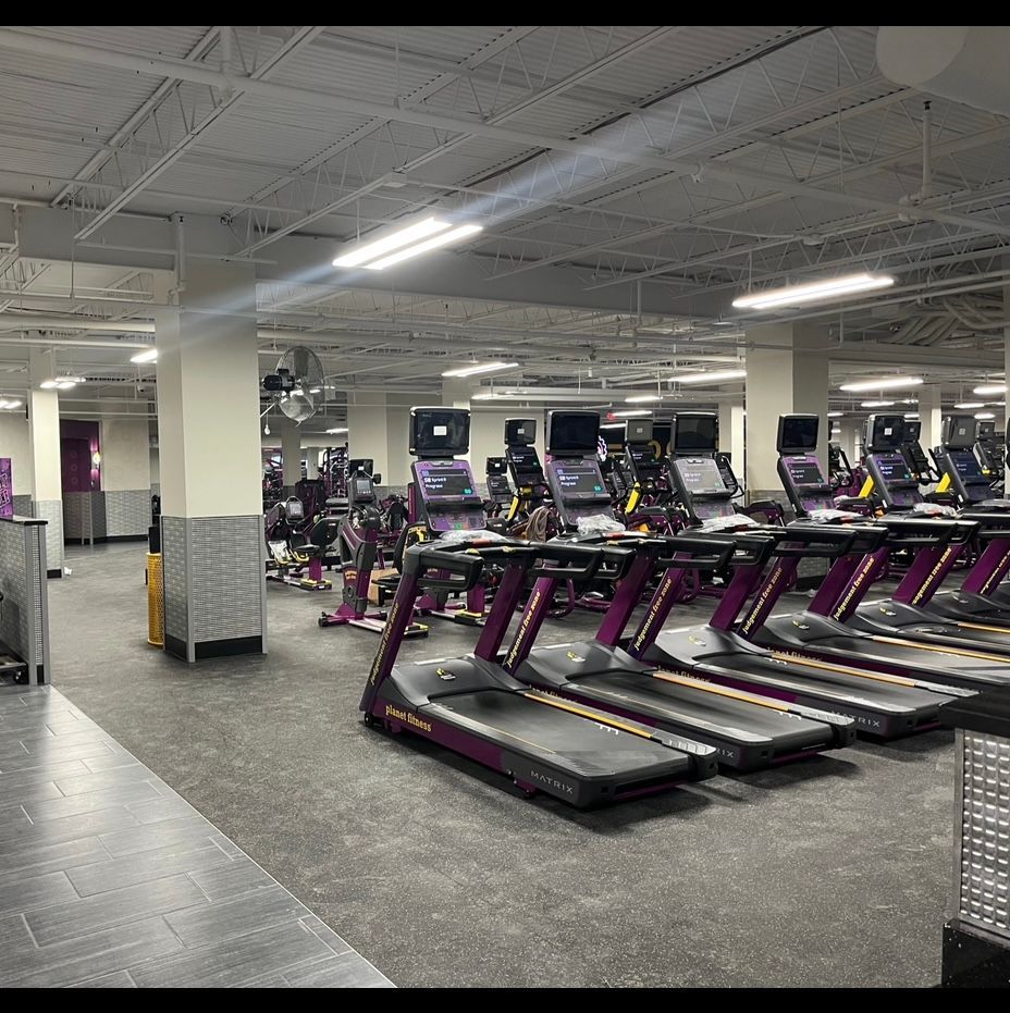 Empty gym with rows of treadmills under bright fluorescent lights and exposed ceiling.