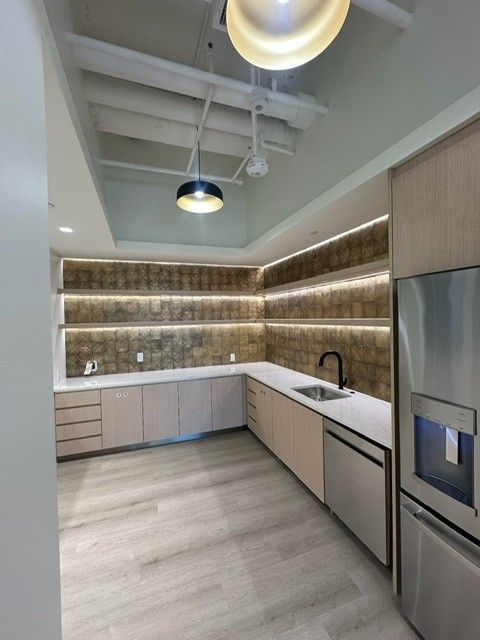 Modern kitchen with light wood cabinets, illuminated shelves, white countertops, and stainless steel appliances