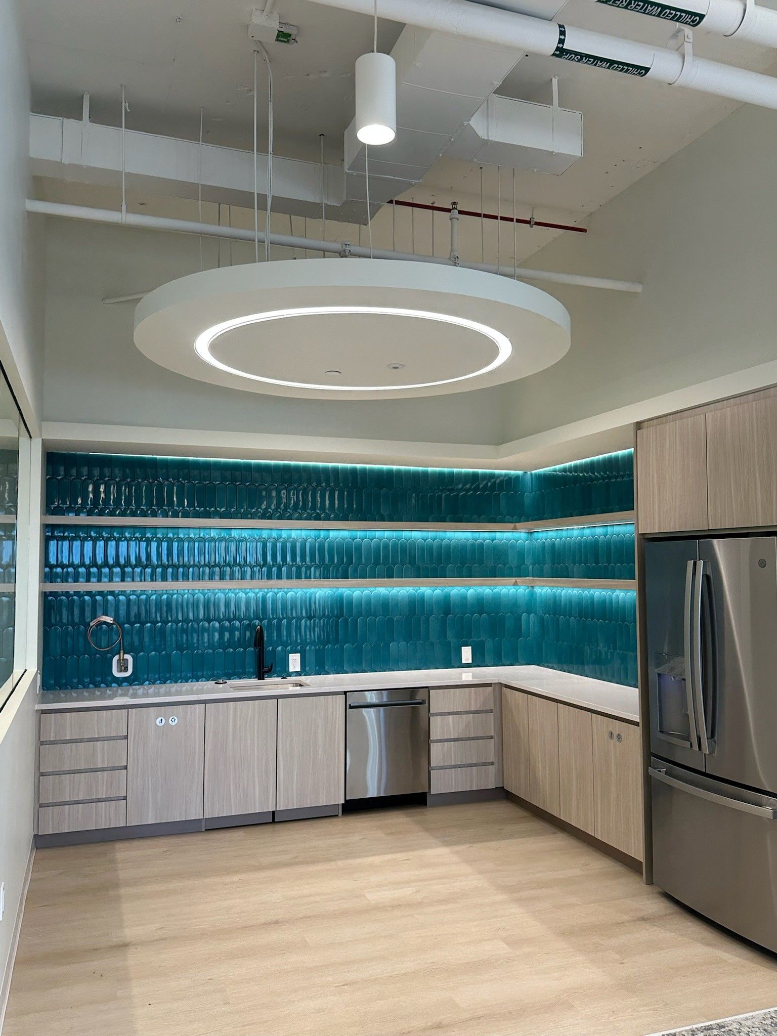 Modern kitchen with light wood cabinets, teal tile backsplash, and a circular ceiling light.