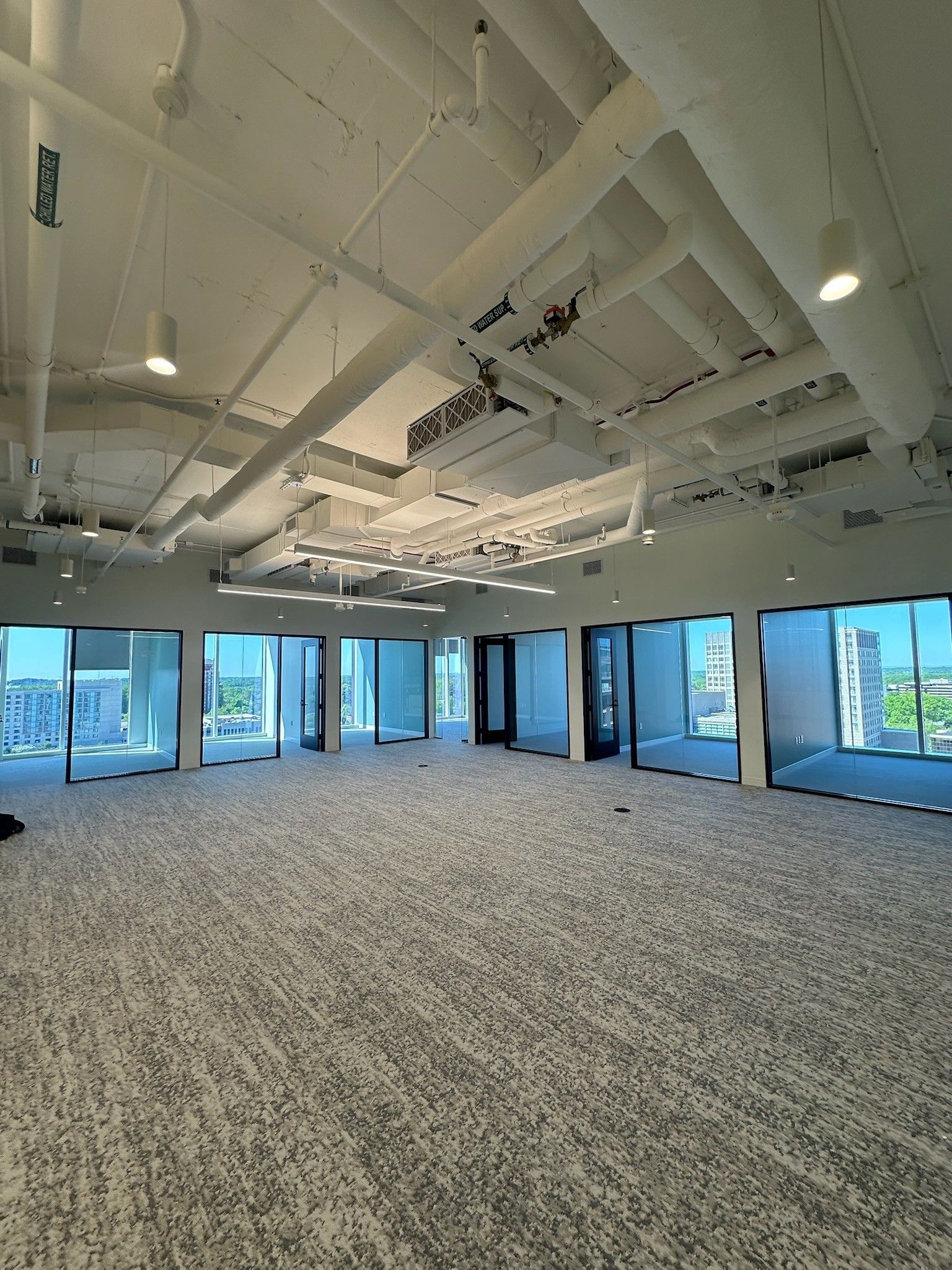 Empty modern office space with carpet, exposed ceiling ducts, and large windows overlooking water