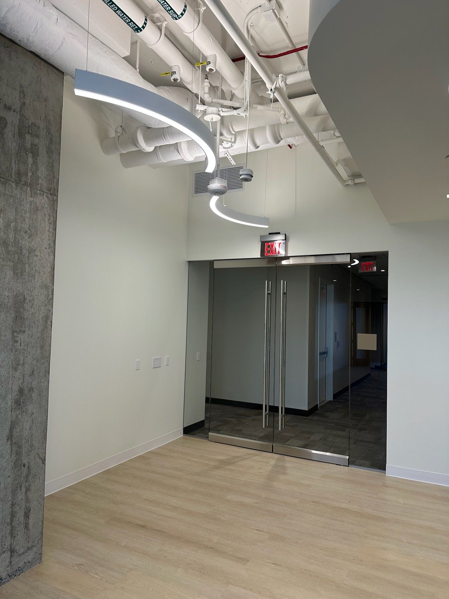 Empty modern office interior with light wood floors, exposed ceiling pipes, and glass double doors.
