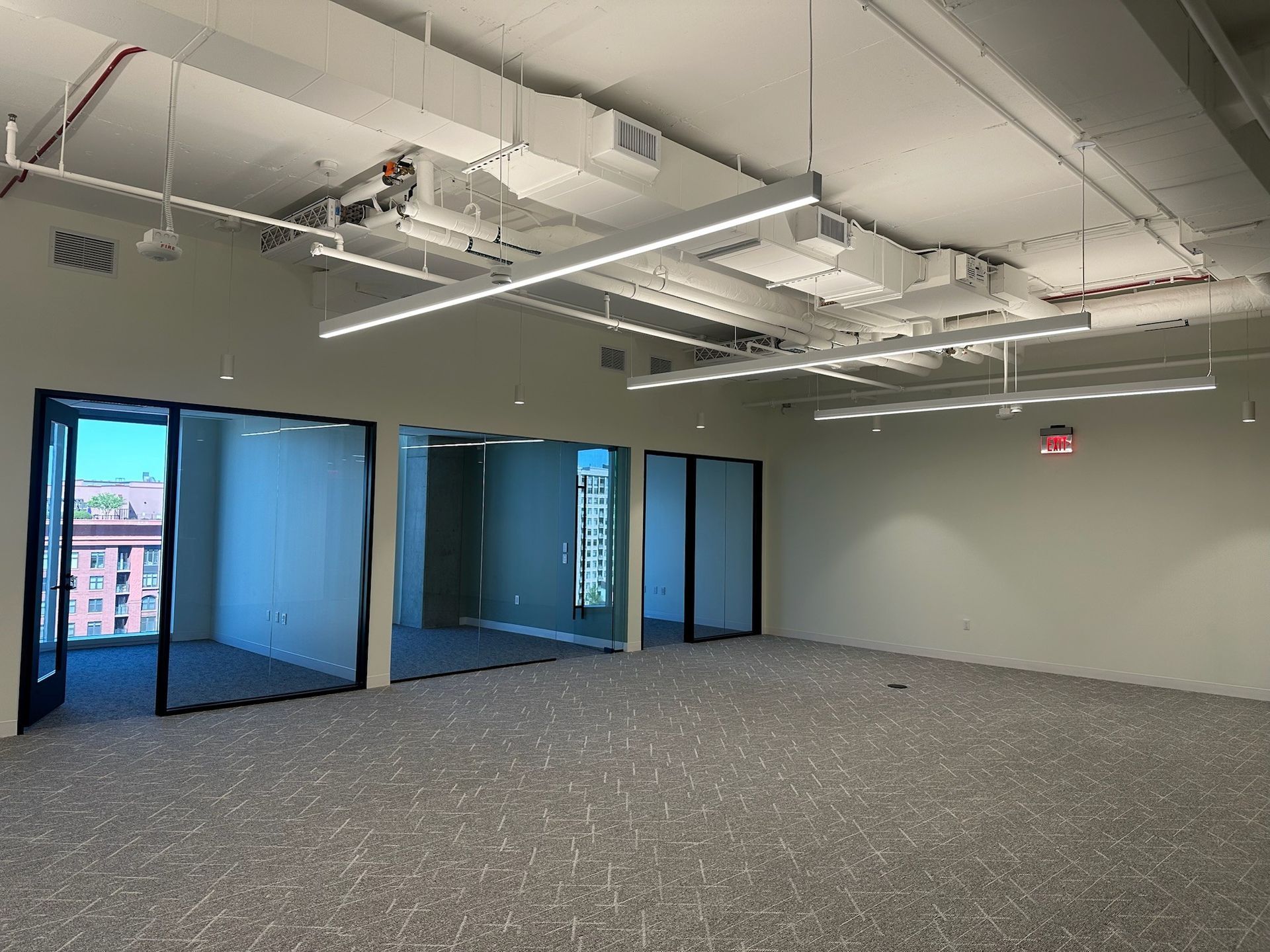 Empty modern office space with carpeted floor, glass partitions, and exposed ceiling fixtures