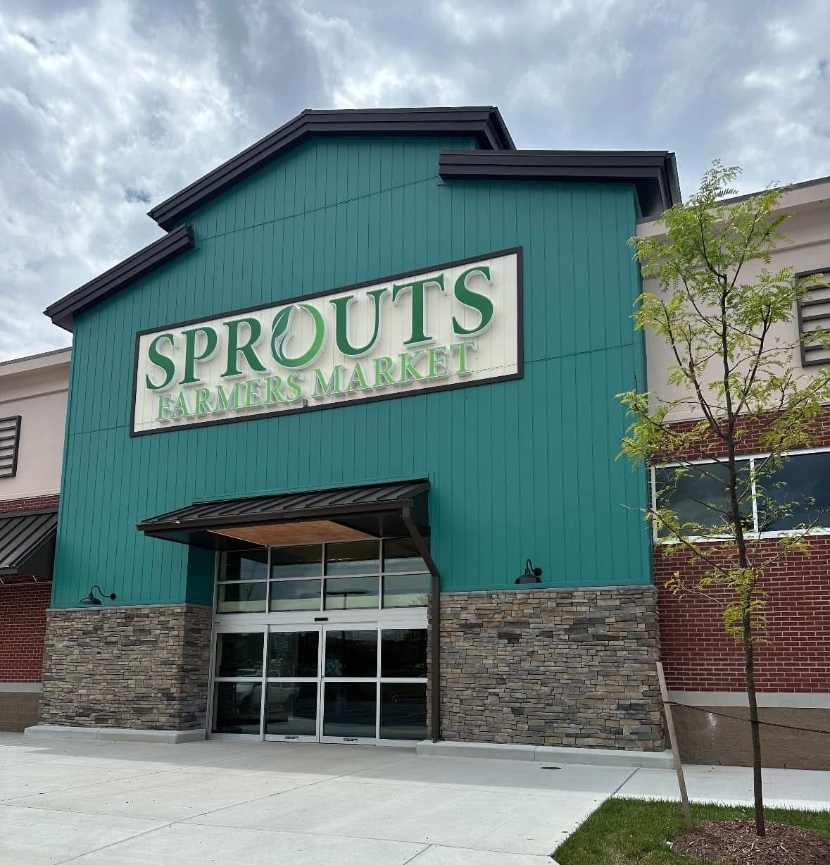 Sprouts grocery store entrance with green facade, stone base, and cloudy sky above.
