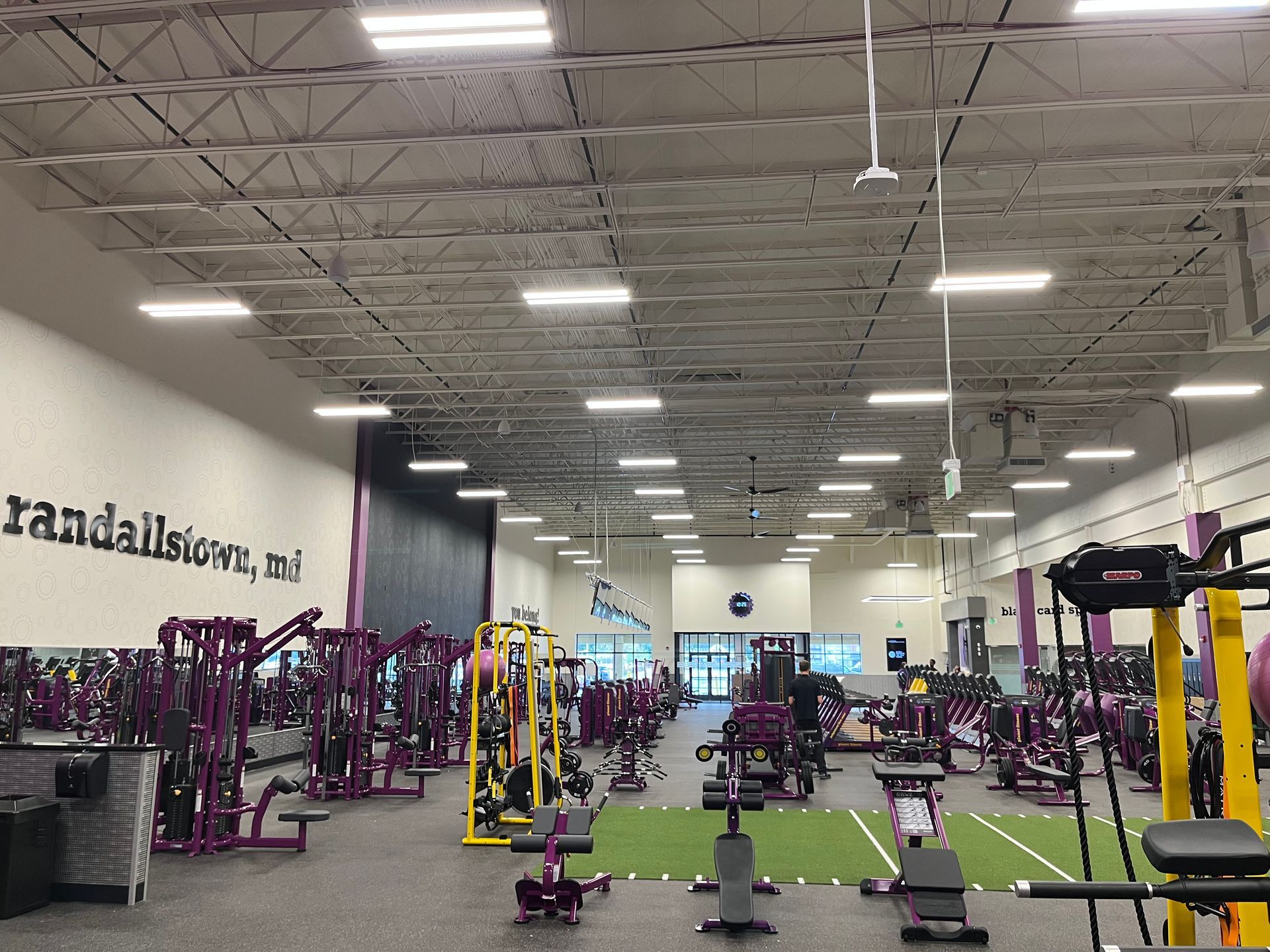 Wide gym interior with rows of purple and yellow exercise machines under high ceiling lights