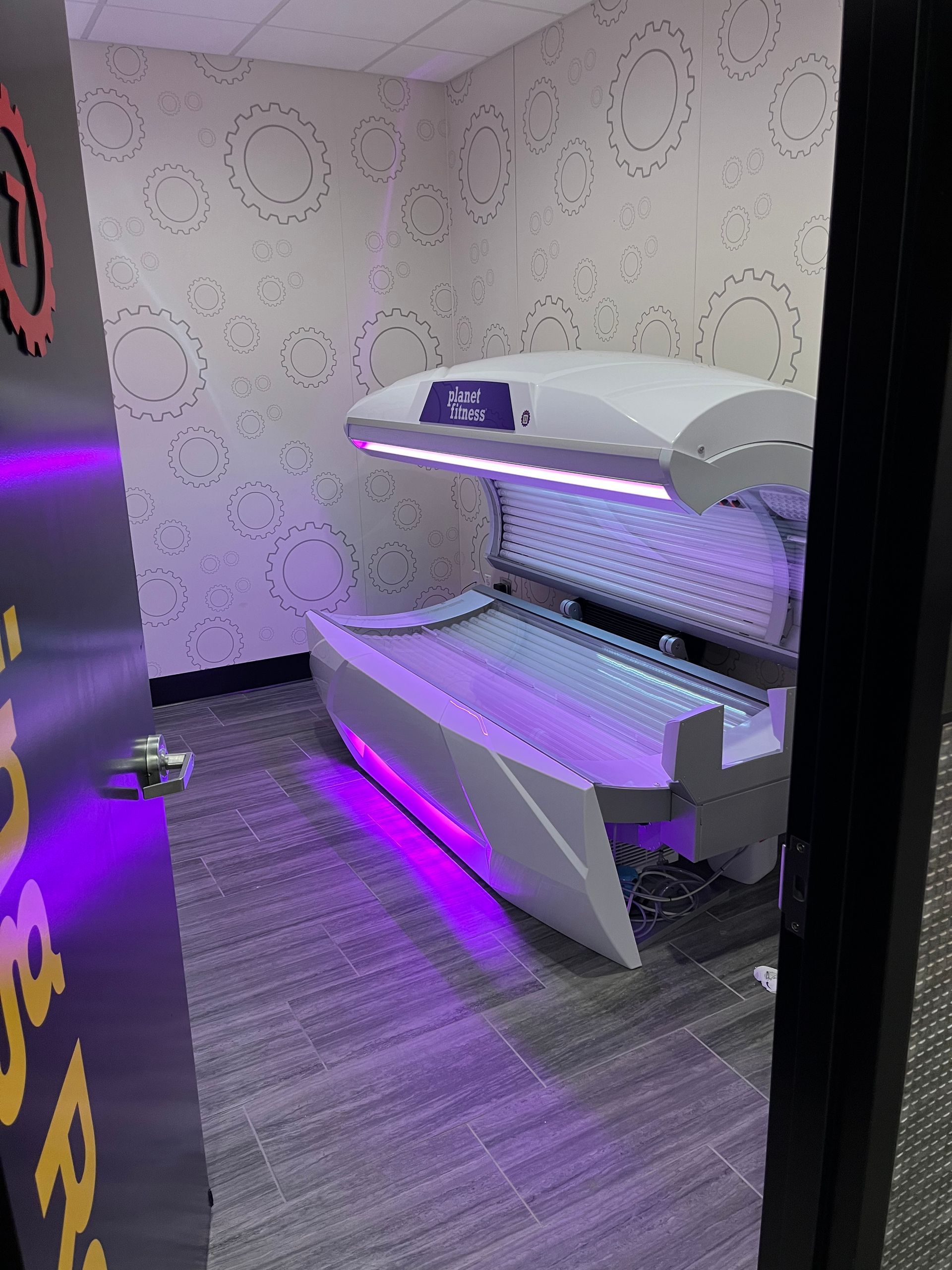 Purple-lit tanning booth in a small room with gray carpet and patterned walls.