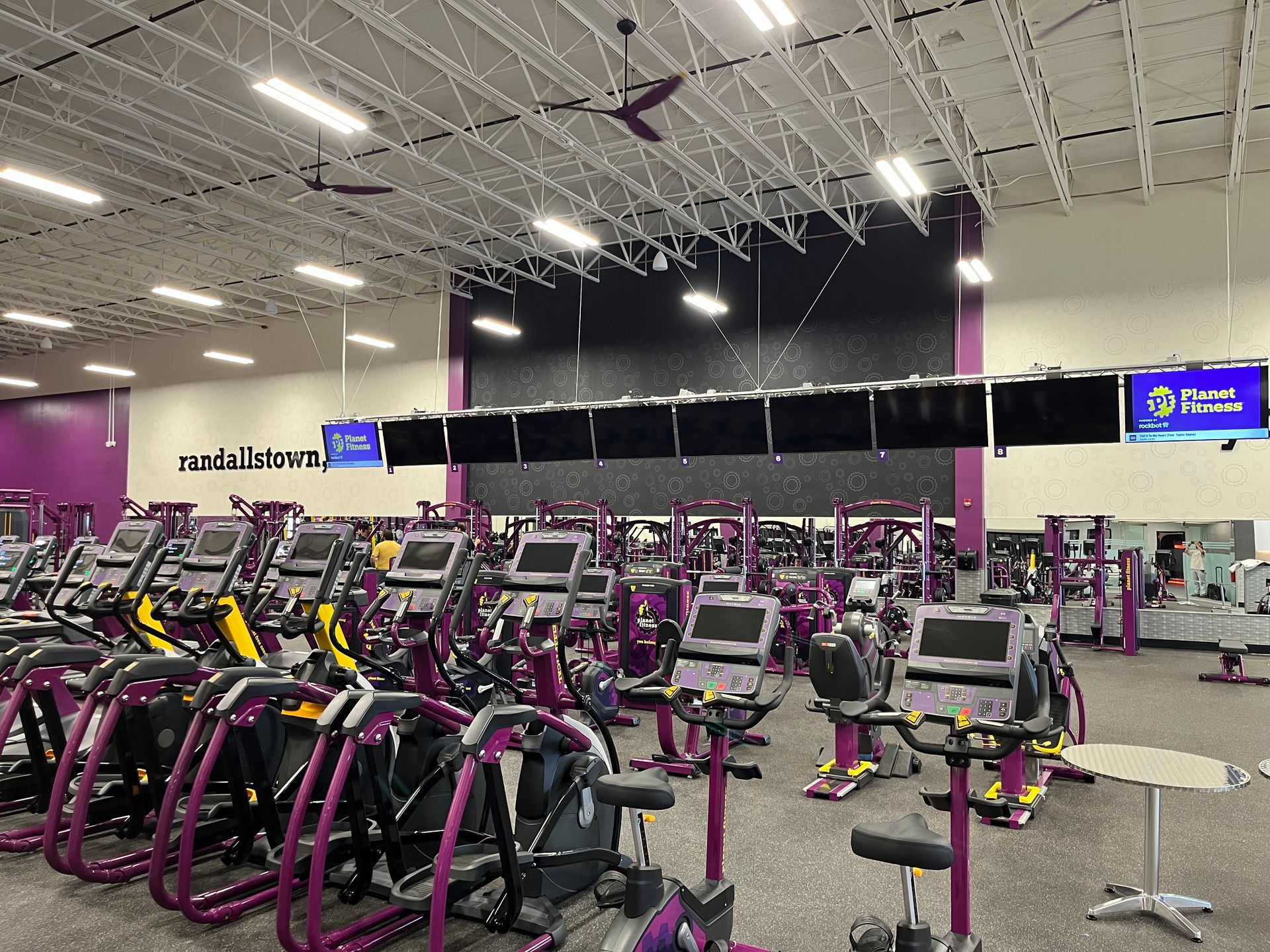 Bright gym with rows of purple and black exercise machines under high white ceiling lights.